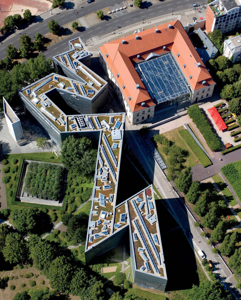 Aerial view of a modern, zigzag-shaped building complex with green roofs next to a traditional building with a red roof and glass atrium, surrounded by trees and roads.
