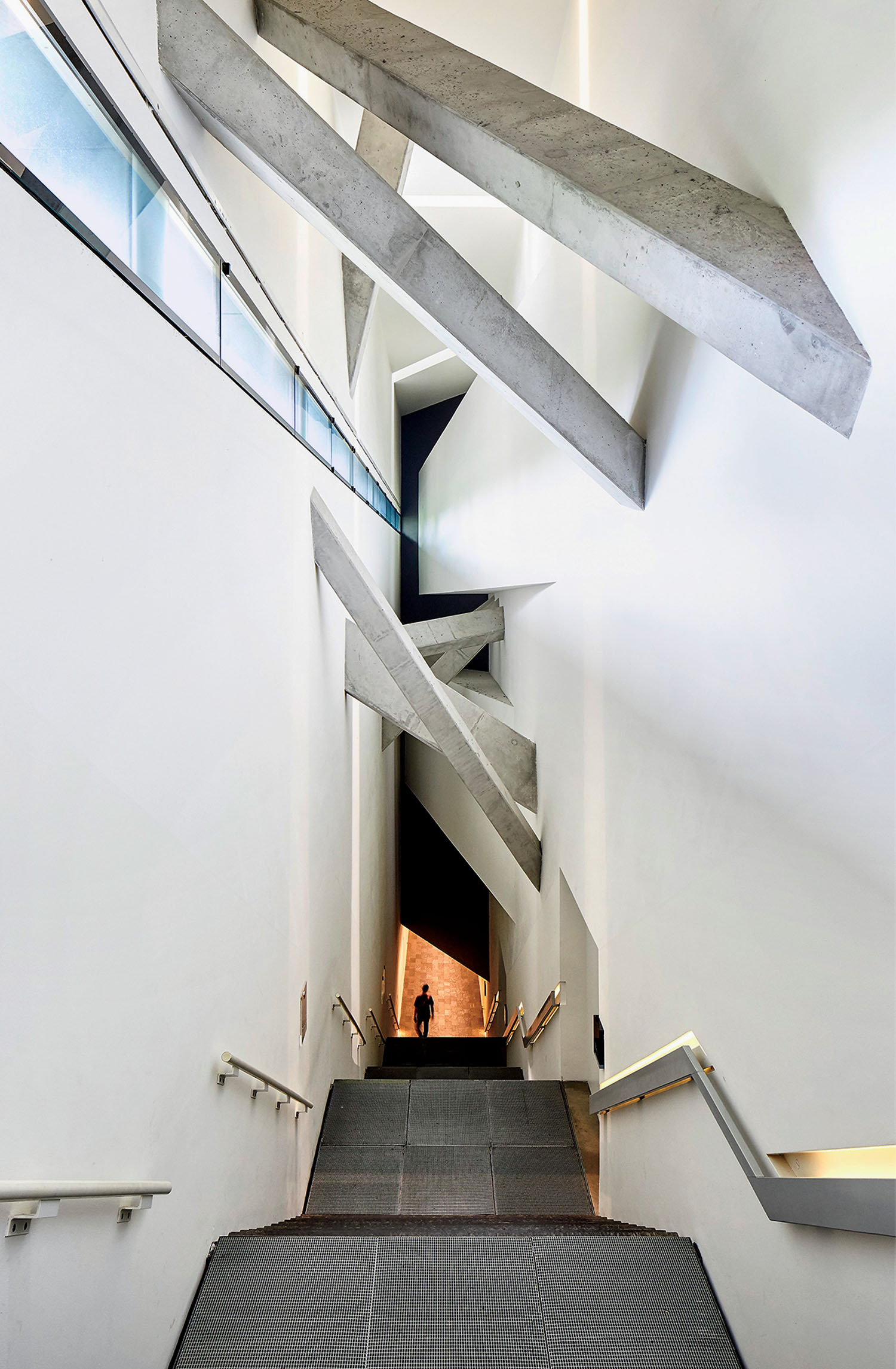 A person stands at the end of a ramp in a modern building with white walls and dramatic angled concrete beams above.