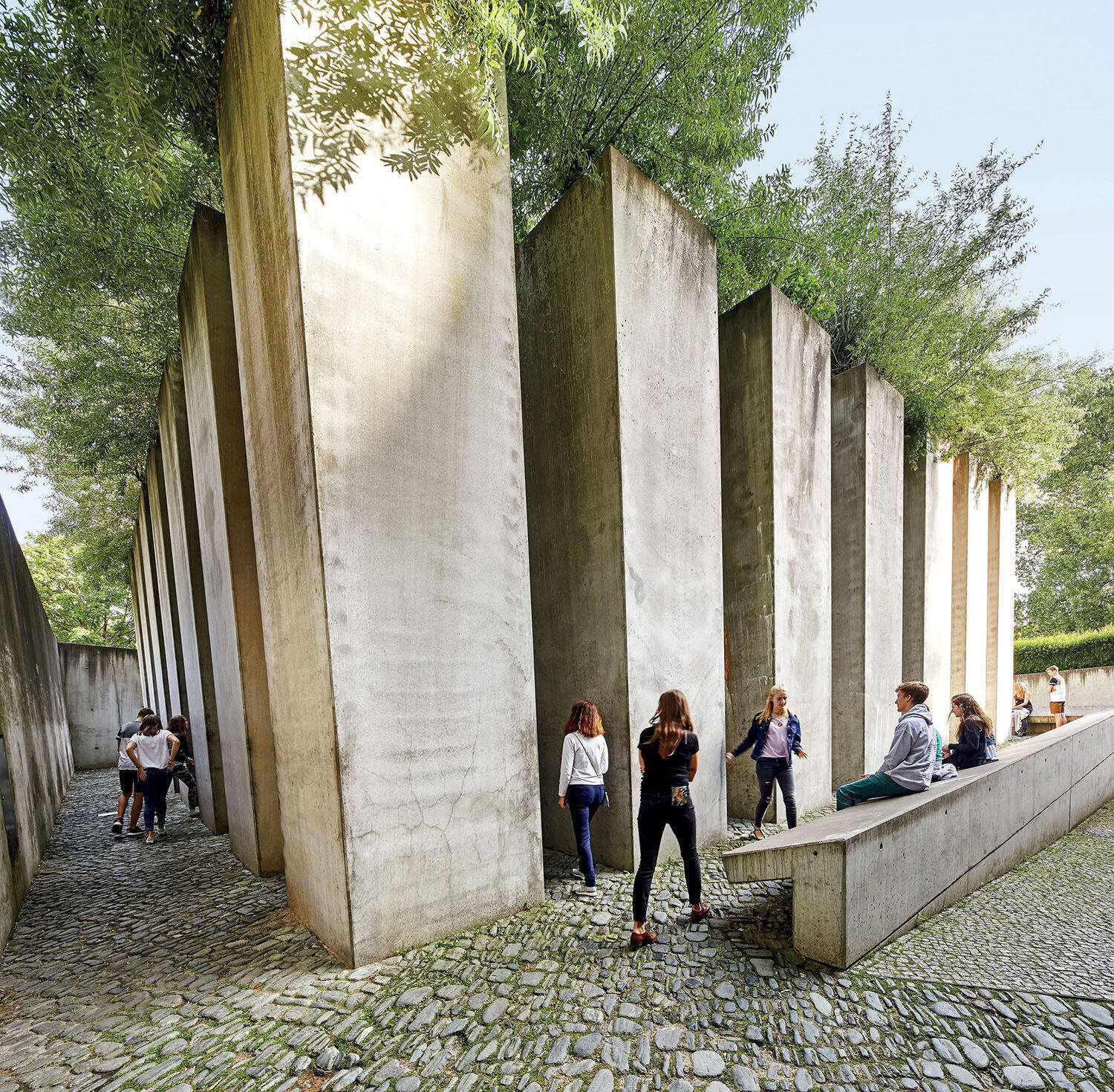 People walk and sit among tall concrete pillars and cobblestone paths, with trees and greenery above, in an outdoor architectural space.