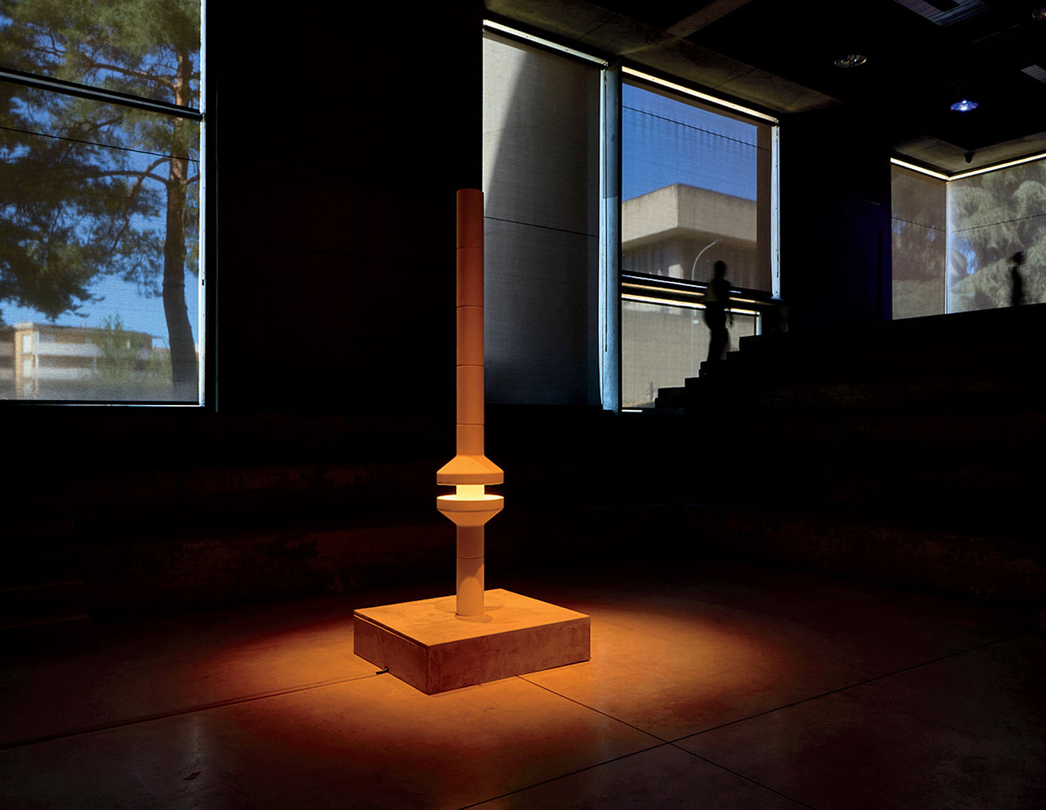 A tall, geometric sculpture stands on a square base, illuminated by a spotlight in a dark room with large windows and silhouetted figures in the background.