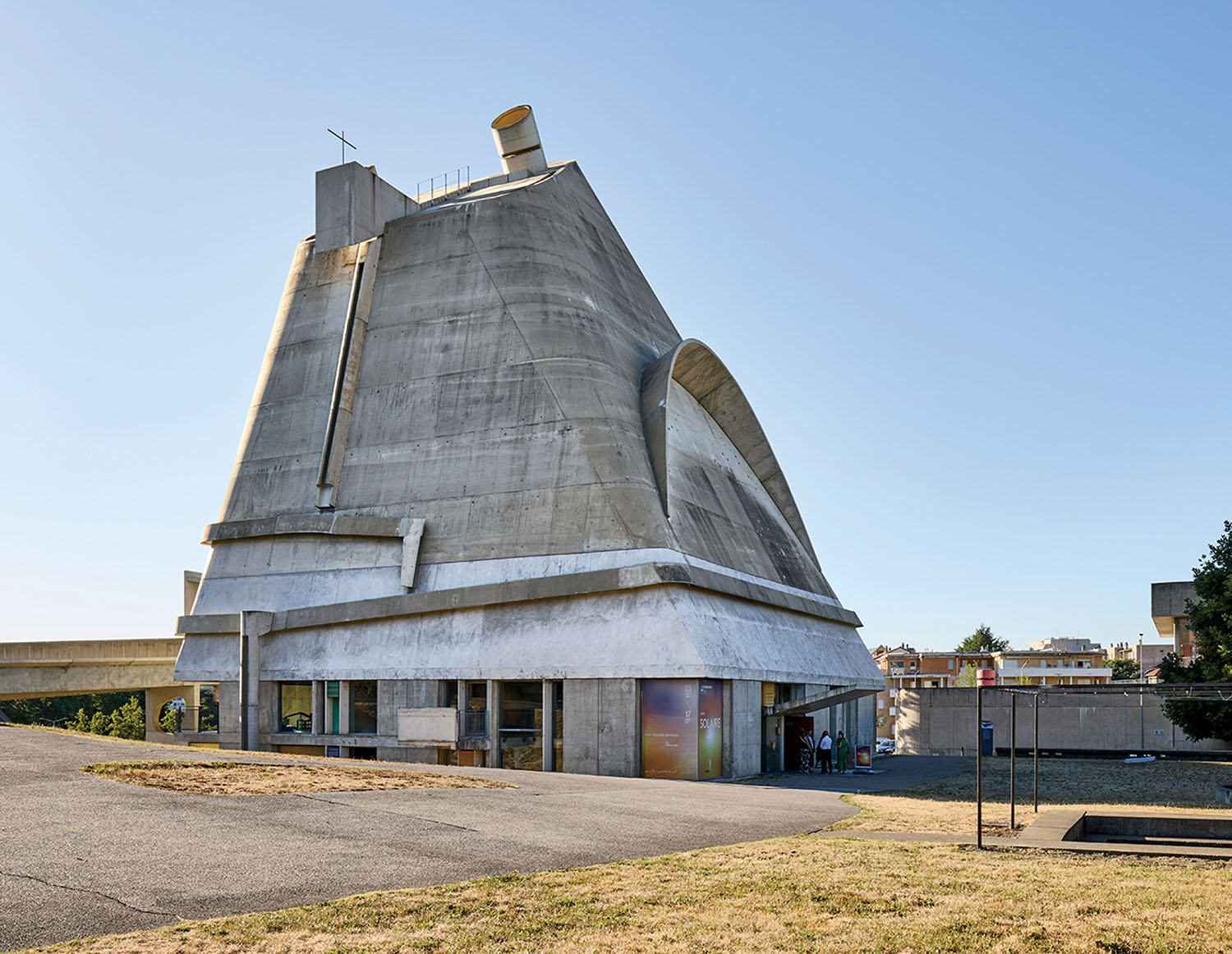 A large, sculptural concrete building with angled walls and a curved roof stands under a clear sky; some people are near the entrance at the base.