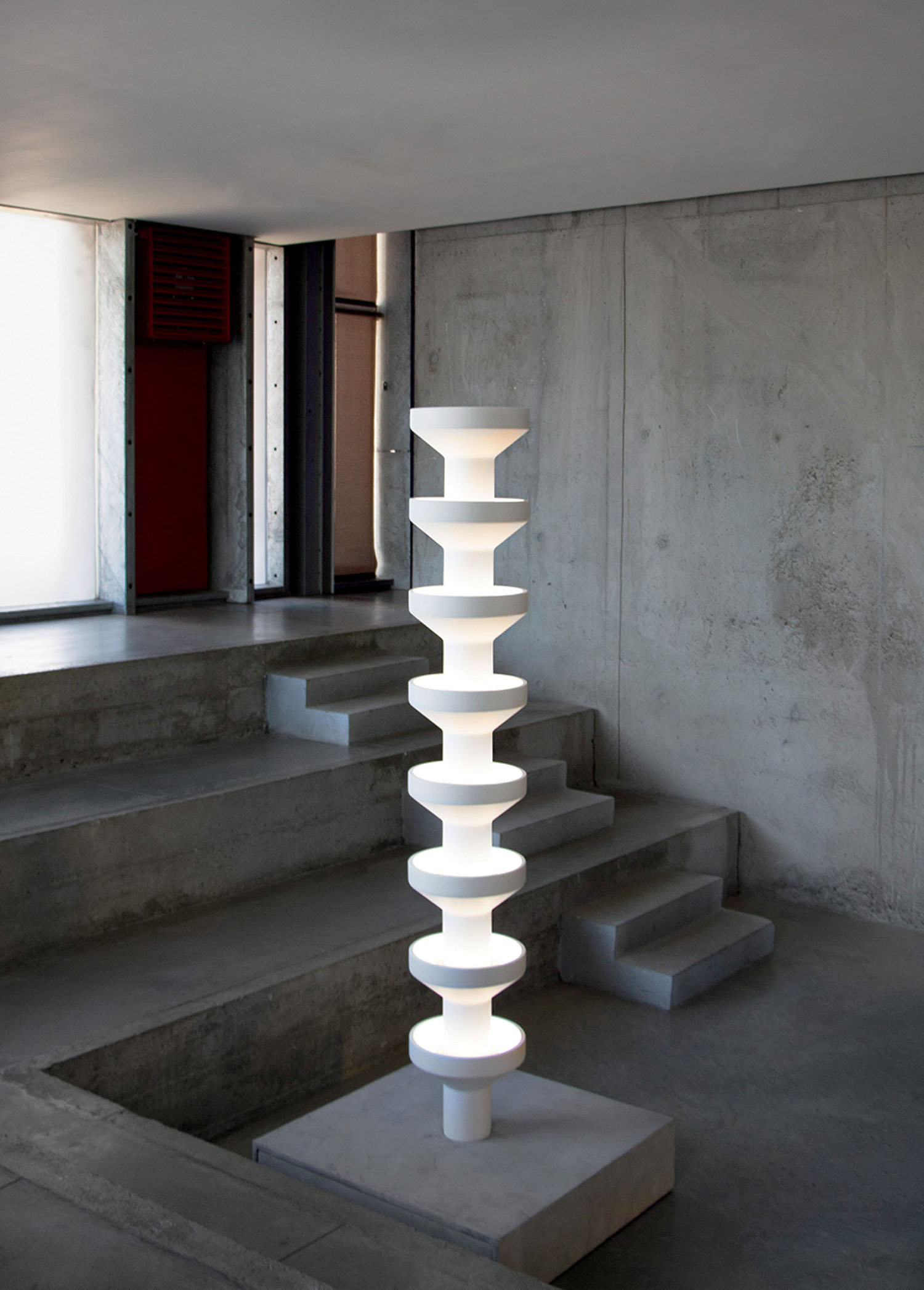 A tall, white, modern sculptural light fixture stands on a concrete floor amid minimalist concrete steps and walls in a contemporary interior space.