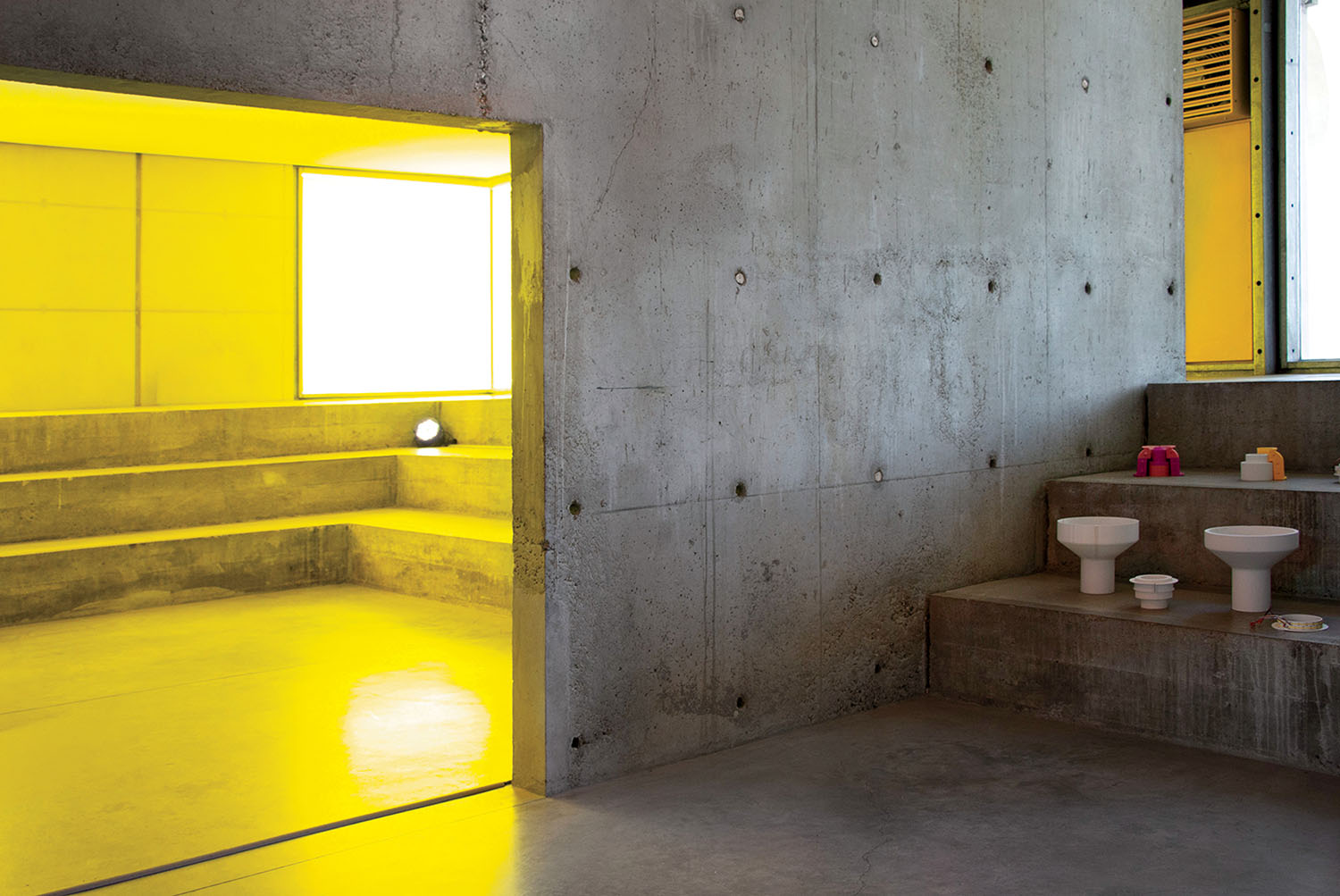 A concrete room with small sinks and toilets on steps; next to it is a bright yellow-lit area with built-in bench seating.