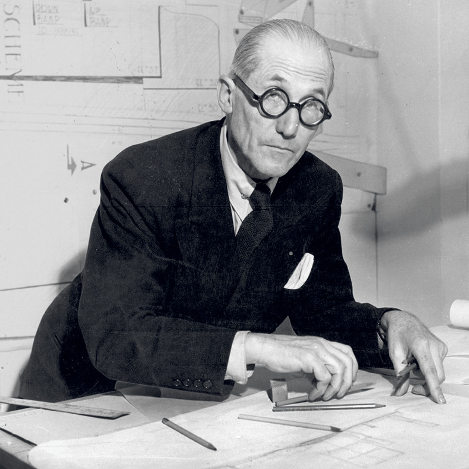 An older man in a suit and round glasses sits at a drafting table with papers and pencils, appearing to work on architectural plans.
