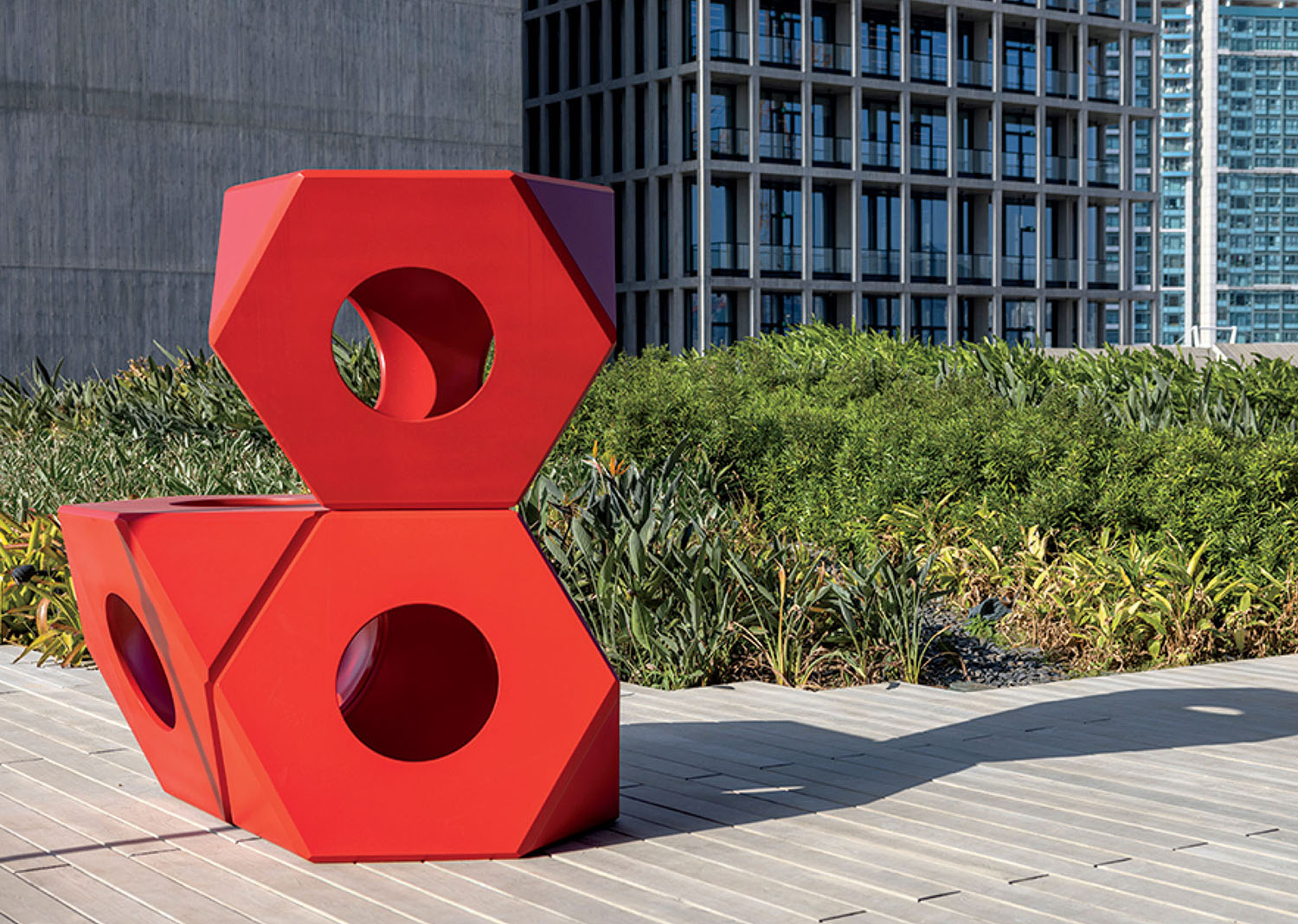 Two large red hexagonal sculptures with circular holes are stacked outdoors on a wooden deck, with green plants and modern buildings in the background.