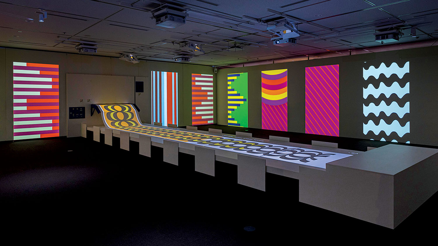 A dark room with seven large, colorful geometric patterns projected on walls and a matching patterned table display in the center.