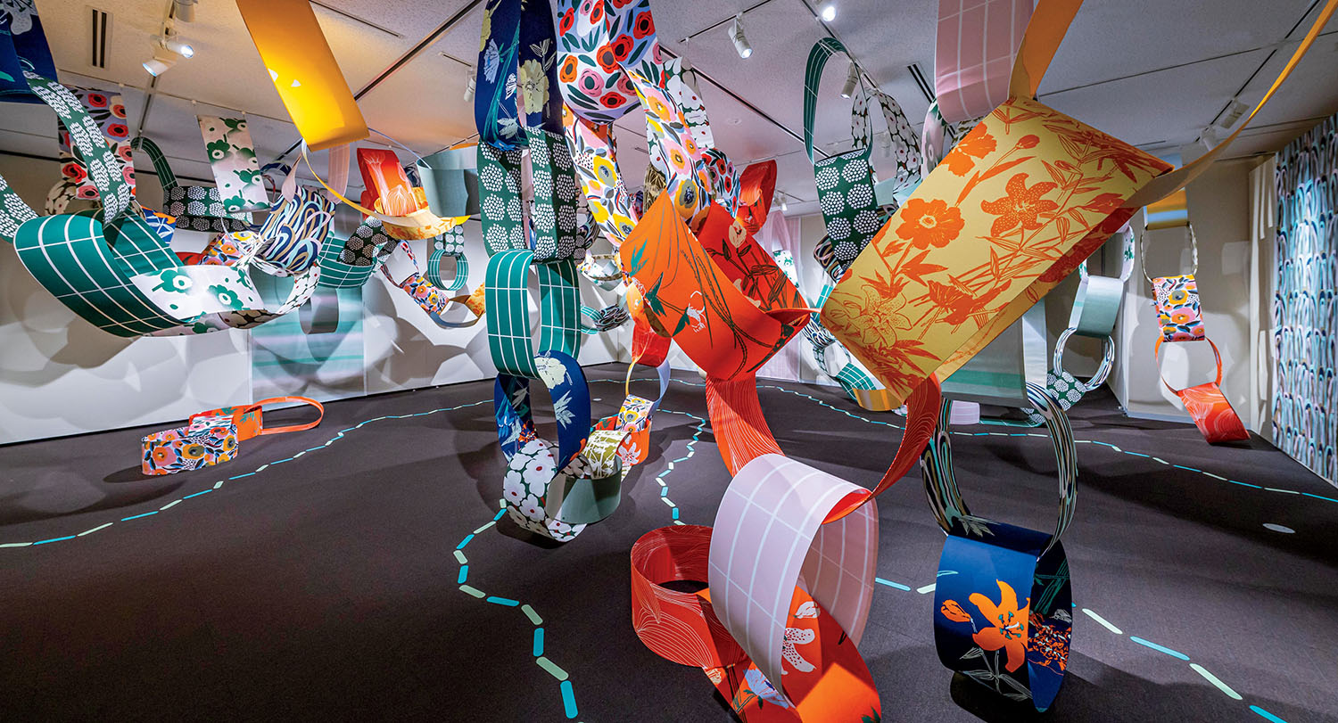 Colorful, patterned paper loops hang from a ceiling in an art installation, creating a dynamic and immersive indoor display with various floral and geometric designs.