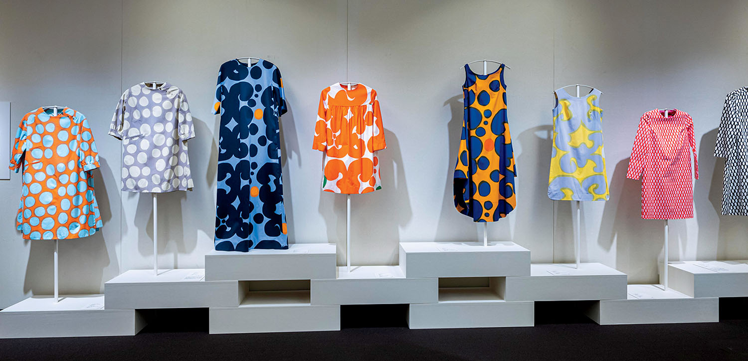 Eight colorful patterned dresses are displayed on white mannequins and pedestals against a plain wall in an exhibition setting.