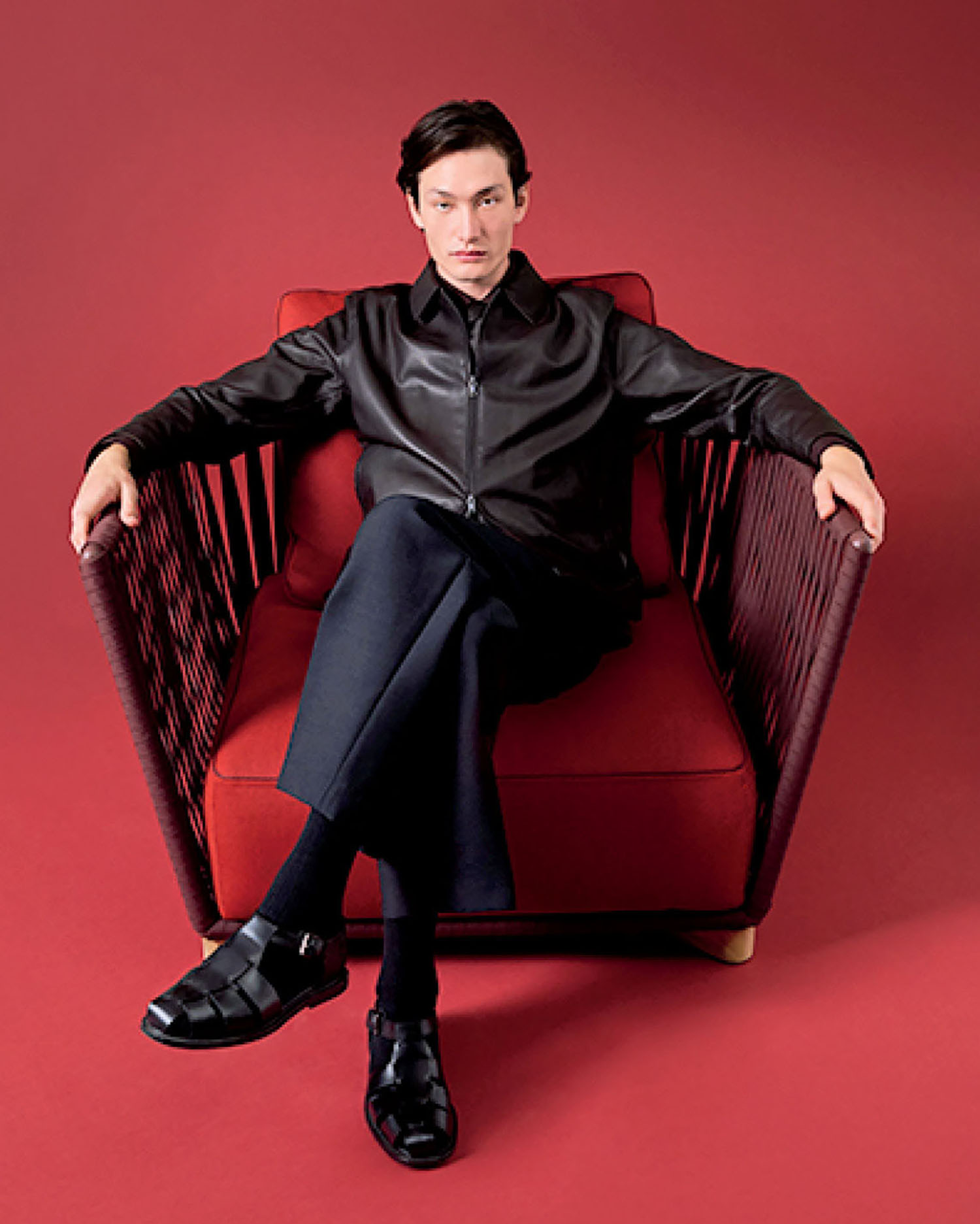 A person in a black jacket and pants sits cross-legged on a red armchair against a solid red background.