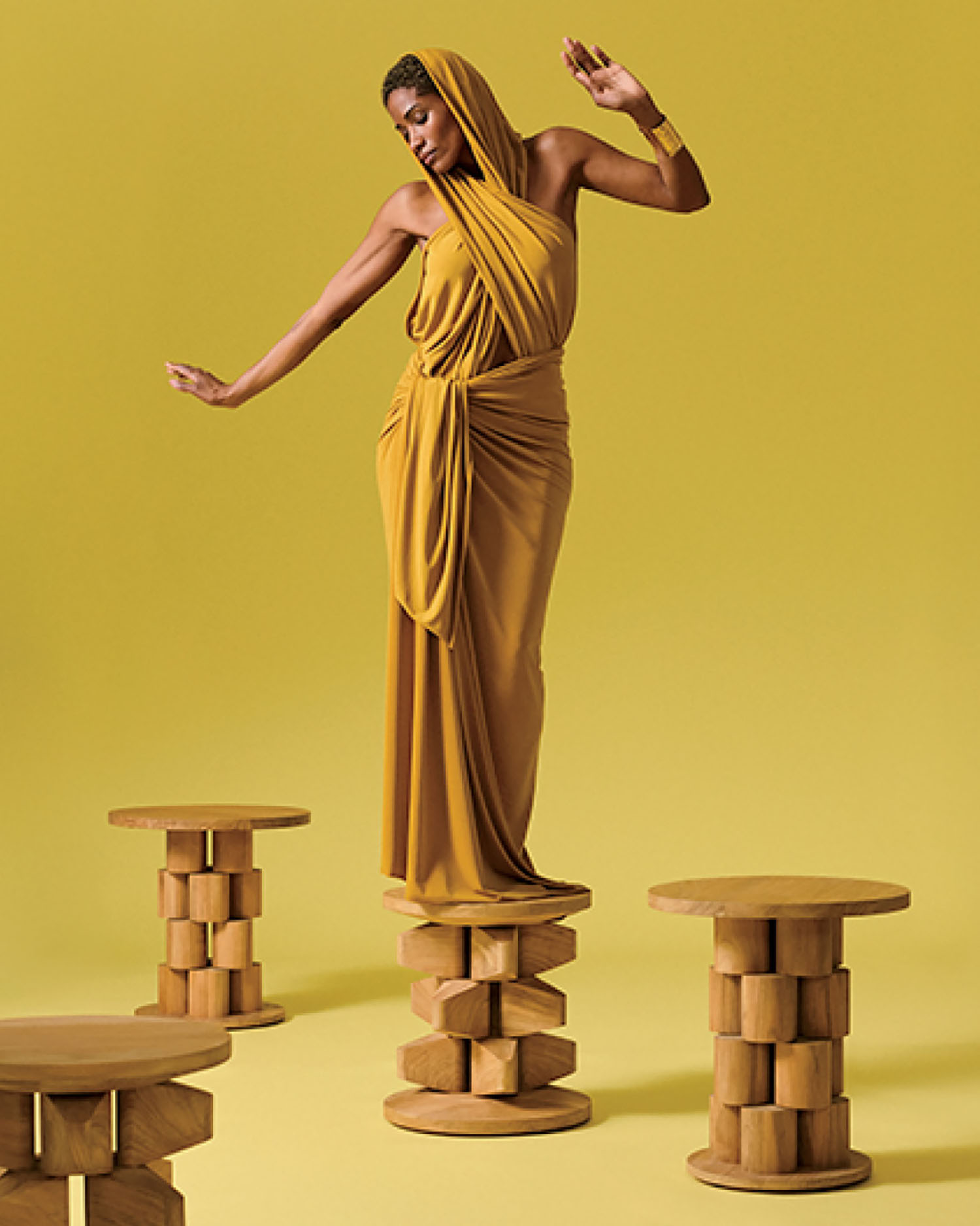 A woman in a gold draped dress stands on a wooden pedestal against a yellow background, with two similar wooden pedestals nearby.