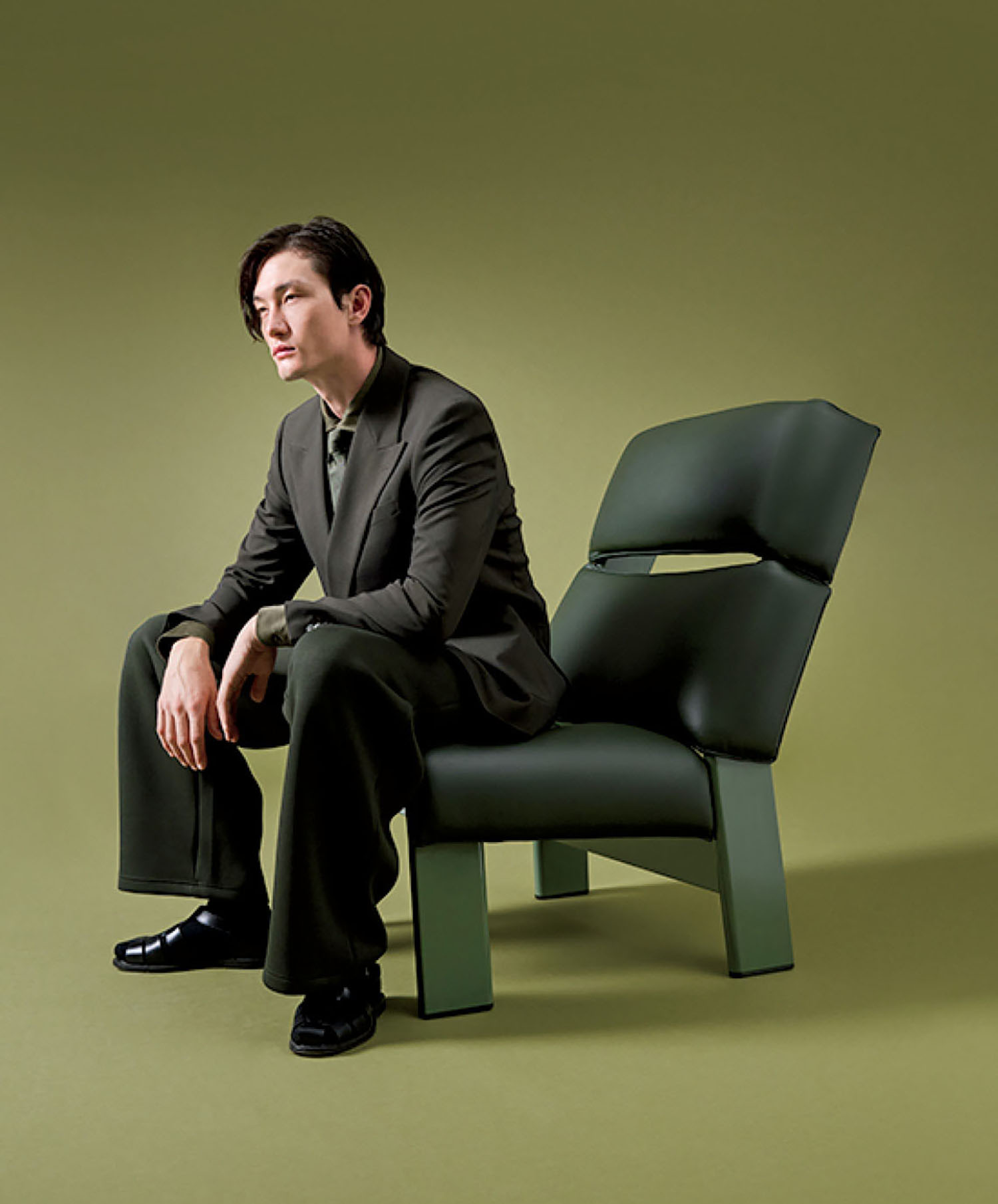 A man in a dark suit sits on a modern, green chair against a matching green background, looking to the left with a neutral expression.