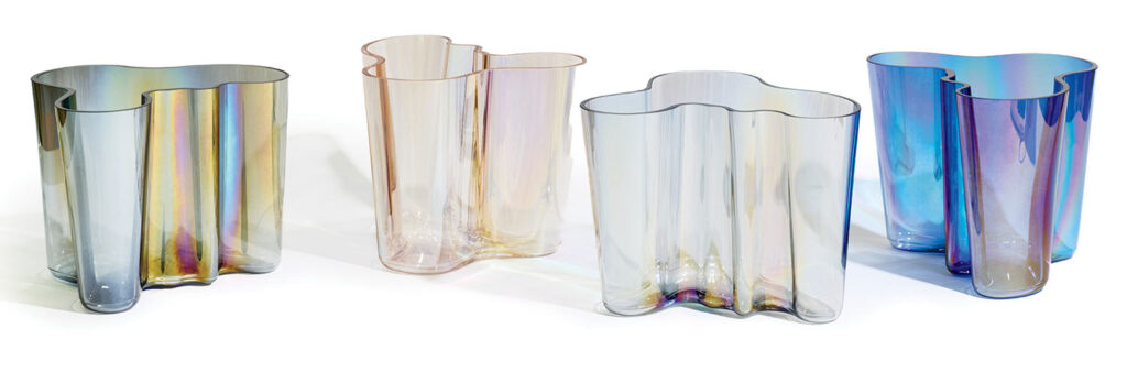 Four wavy-edged, translucent glass vases in pastel yellow, pale pink, light gray, and iridescent blue are lined up against a white background.