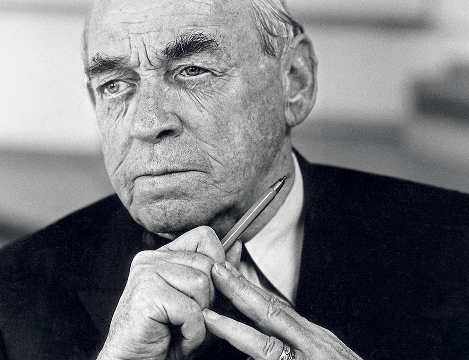 Older man in a suit holds a pen near his face, looking thoughtful with a serious expression.