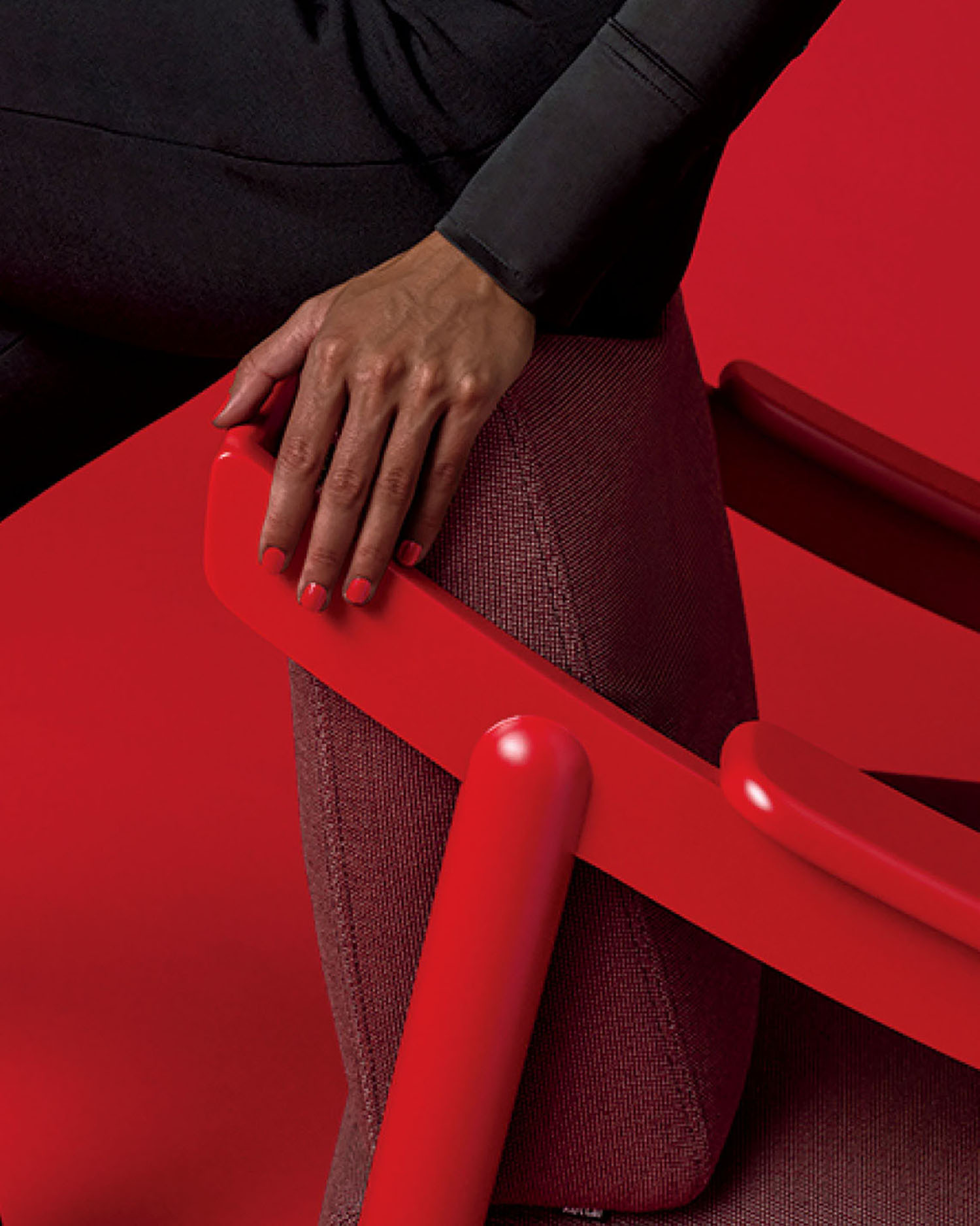 A person with red nail polish rests their hand on the red armrest of a modern chair against a red background.