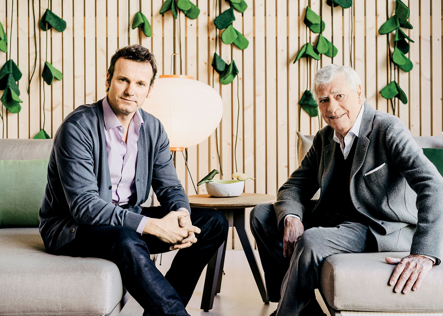 Two men in suits sit on modern sofas in a well-lit room with wooden paneling and green leaf decorations on the wall behind them.