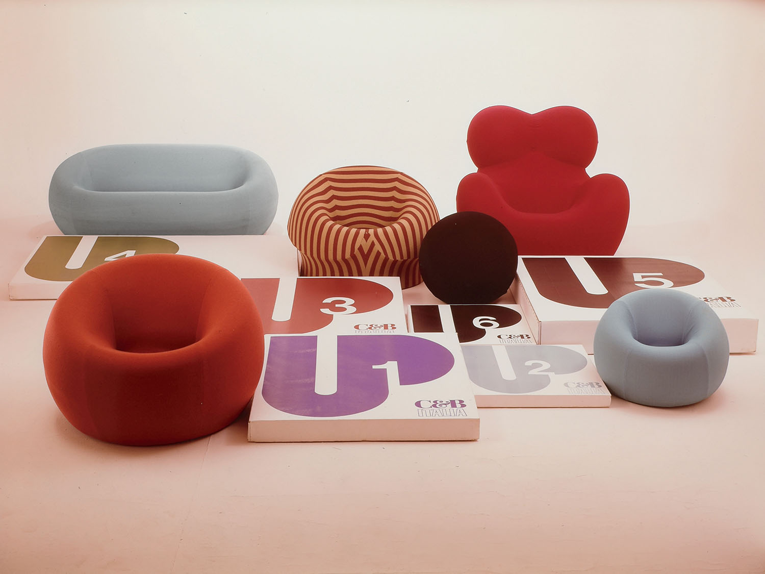 A group of colorful, modern chairs and round pillows are arranged behind large white boxes with bold numbers and letters on their covers, set against a plain background.
