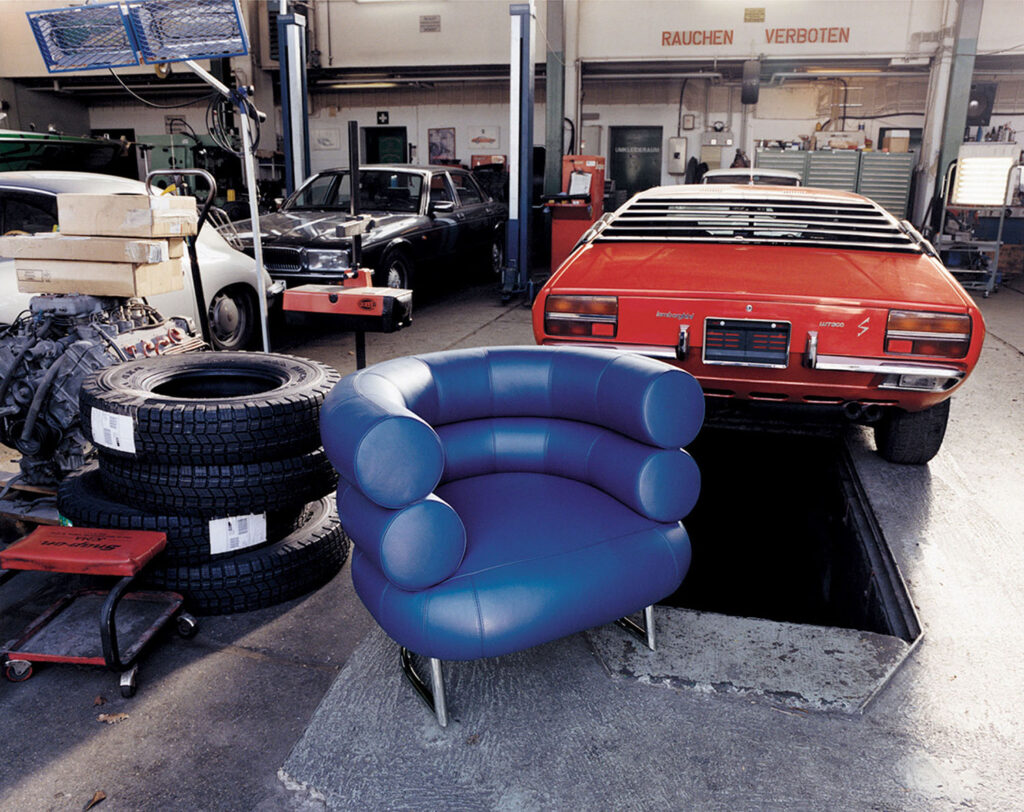 A blue, modern armchair is placed in a car repair shop, in front of a red vintage car over a maintenance pit, with stacked tires nearby.