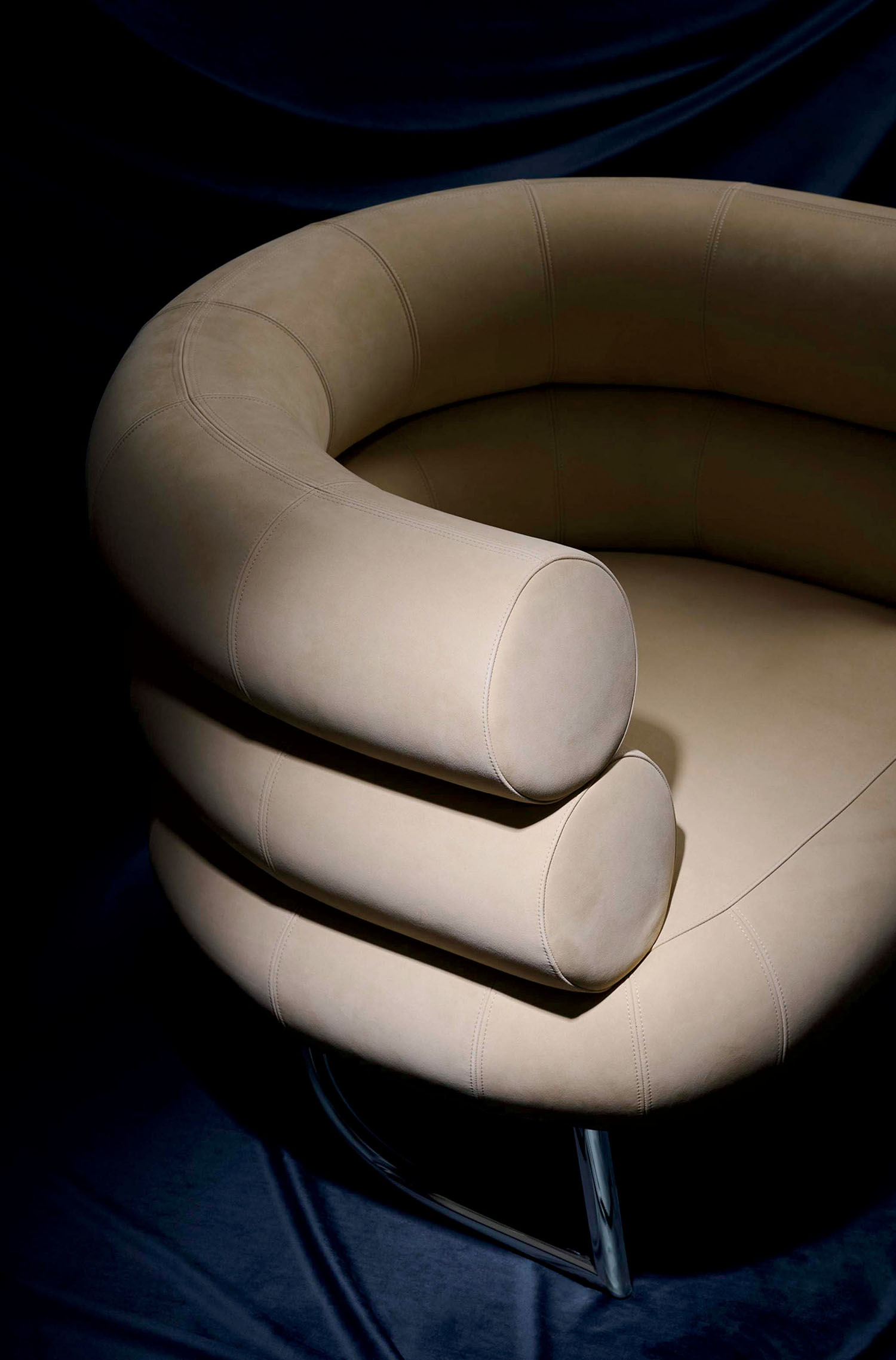A modern beige armchair with rounded, layered armrests and metal legs, set against a dark fabric background.