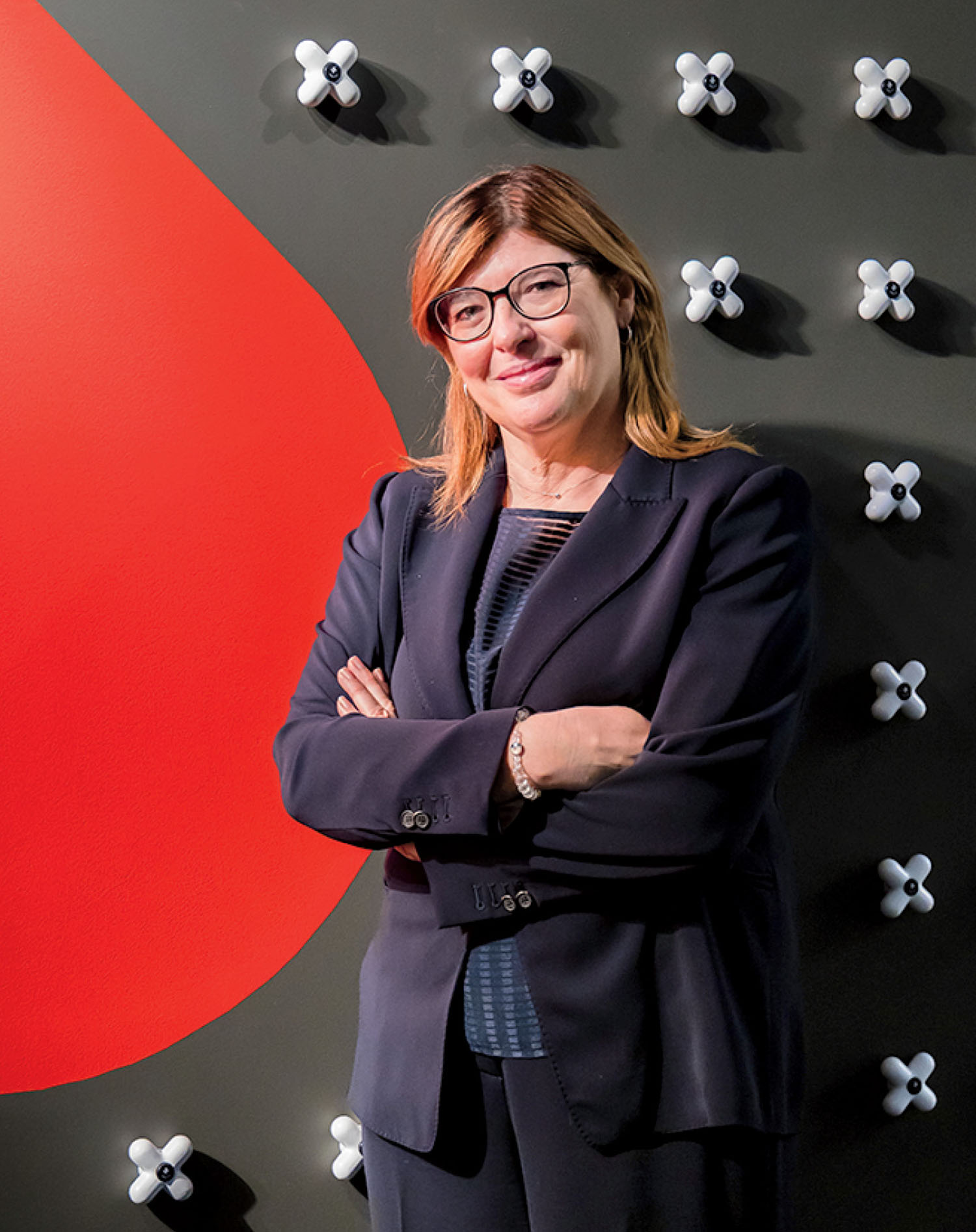A woman in a dark suit stands with arms crossed in front of a wall with a red shape and white decorative elements.
