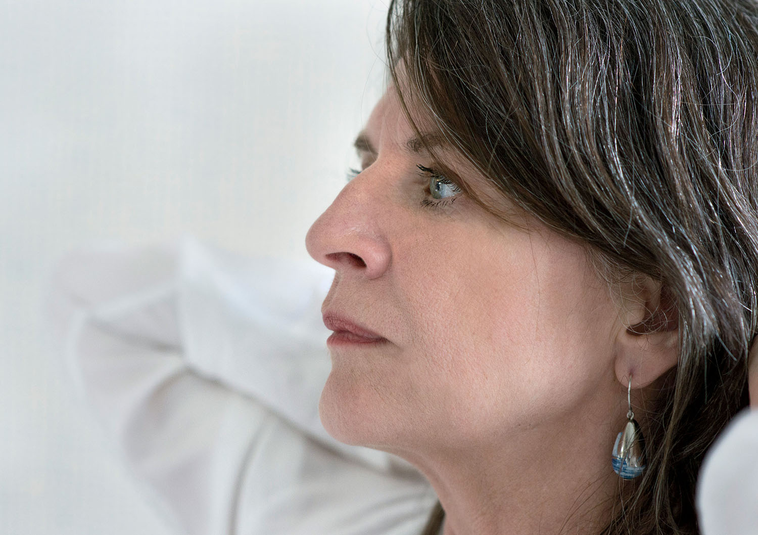 Woman with medium-length brown hair in profile, wearing a white top and a blue teardrop earring, looking thoughtfully into the distance.
