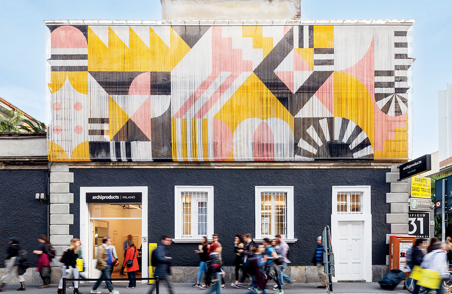 A building with a black lower facade and geometric mural in pink, yellow, and white on the upper facade. People walk by the entrance on a busy street.