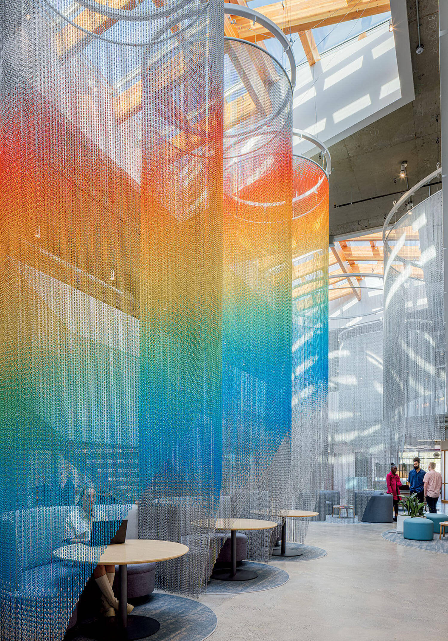 Colorful mesh curtains hang in a modern, open office space with high ceilings, skylights, and people working and conversing at tables.