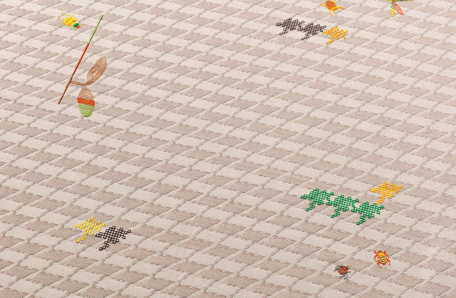 A beige textured fabric with a checkered pattern features small, colorful embroidered designs, including leaves and abstract shapes, scattered across the surface.