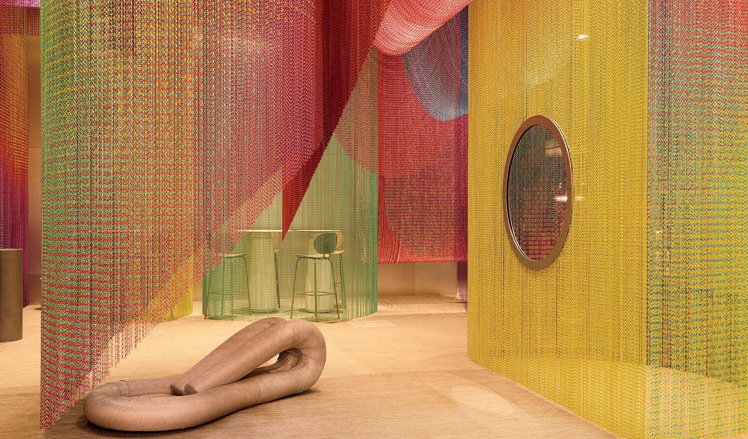 Colorful chain curtains hang in a room with a circular window, a barstool by a glass table, and a large soft sculpture on the floor.