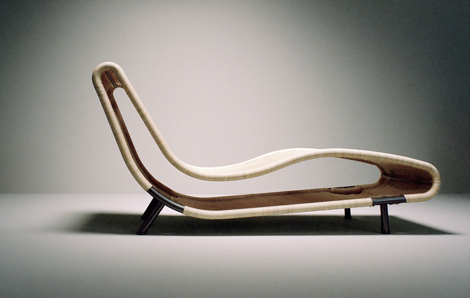 A modern chaise lounge chair with a wavy, minimalist design in light and dark brown tones, set against a plain, neutral background.