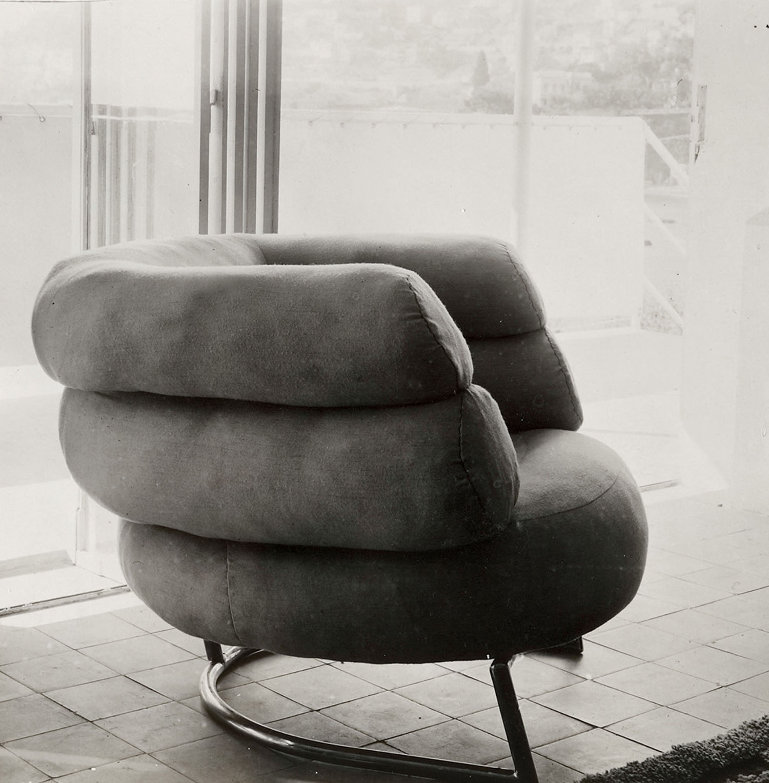 Plush, rounded armchair with a tubular metal base sits on a tiled floor near large windows overlooking an urban landscape.