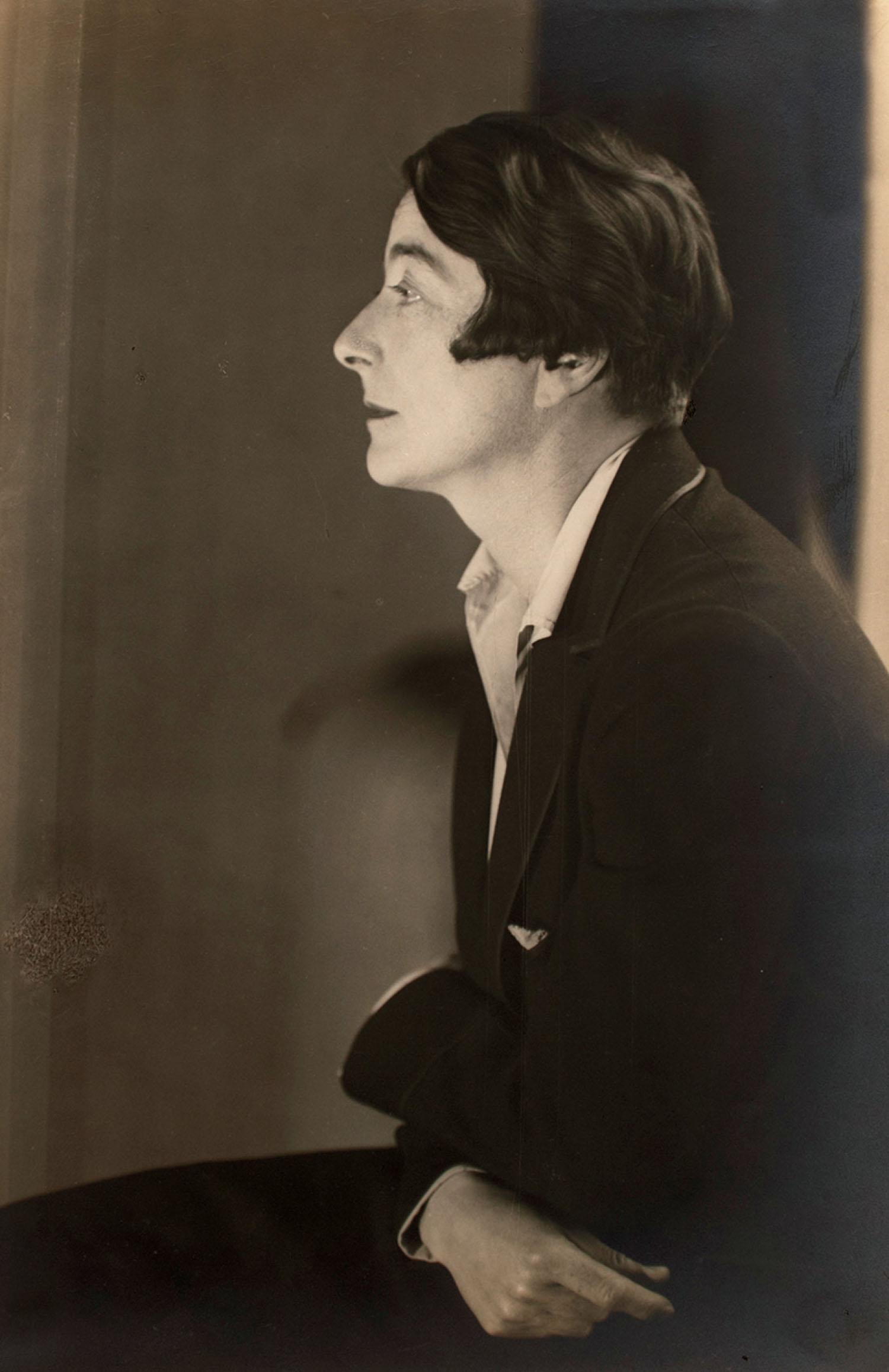 Black and white photo of a woman in profile with short dark hair, wearing a dark jacket and light shirt, sitting and looking to the left.