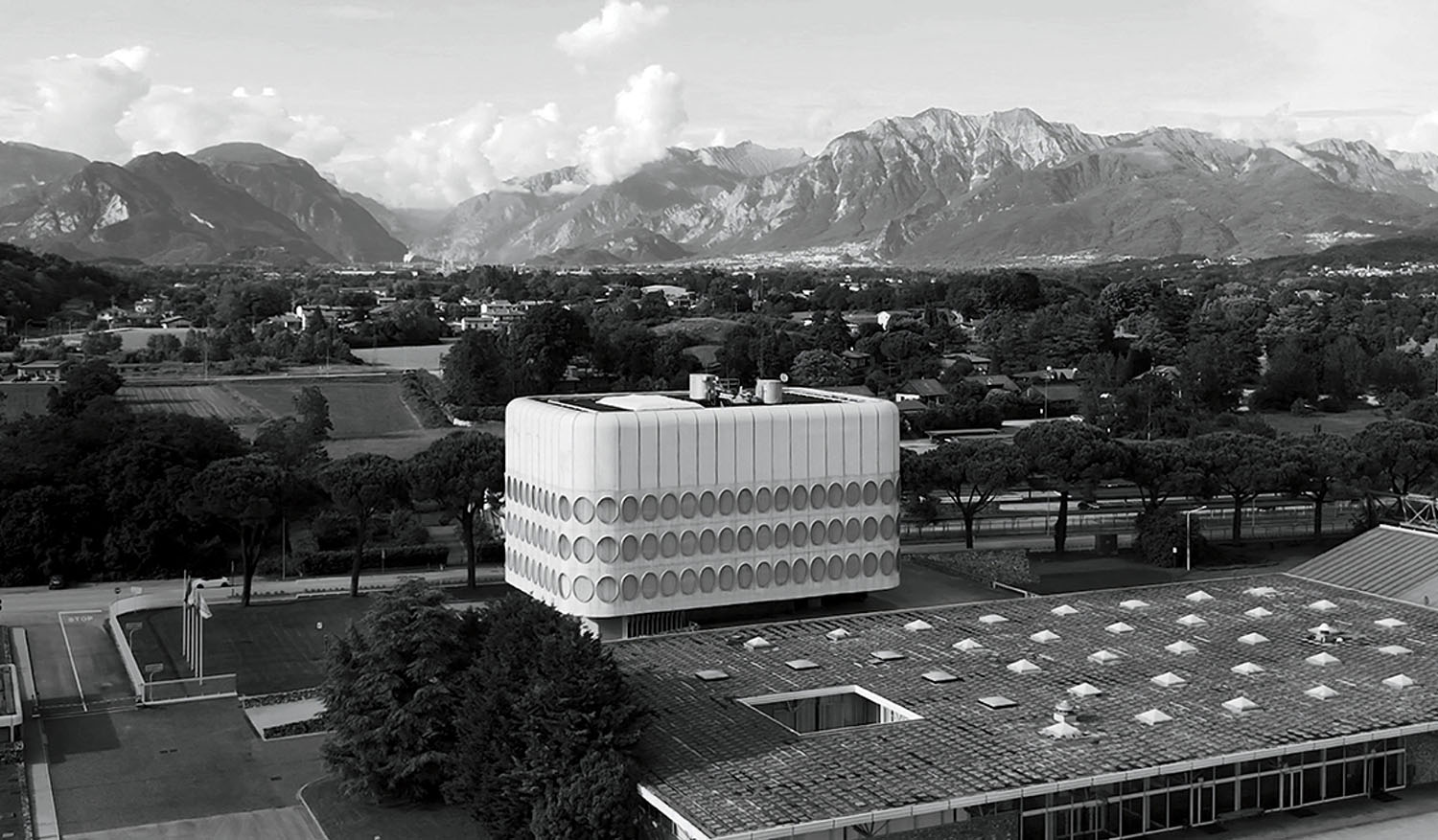 A modern cubic building with circular patterns stands in a semi-rural area with mountains in the background and scattered trees surrounding the site.