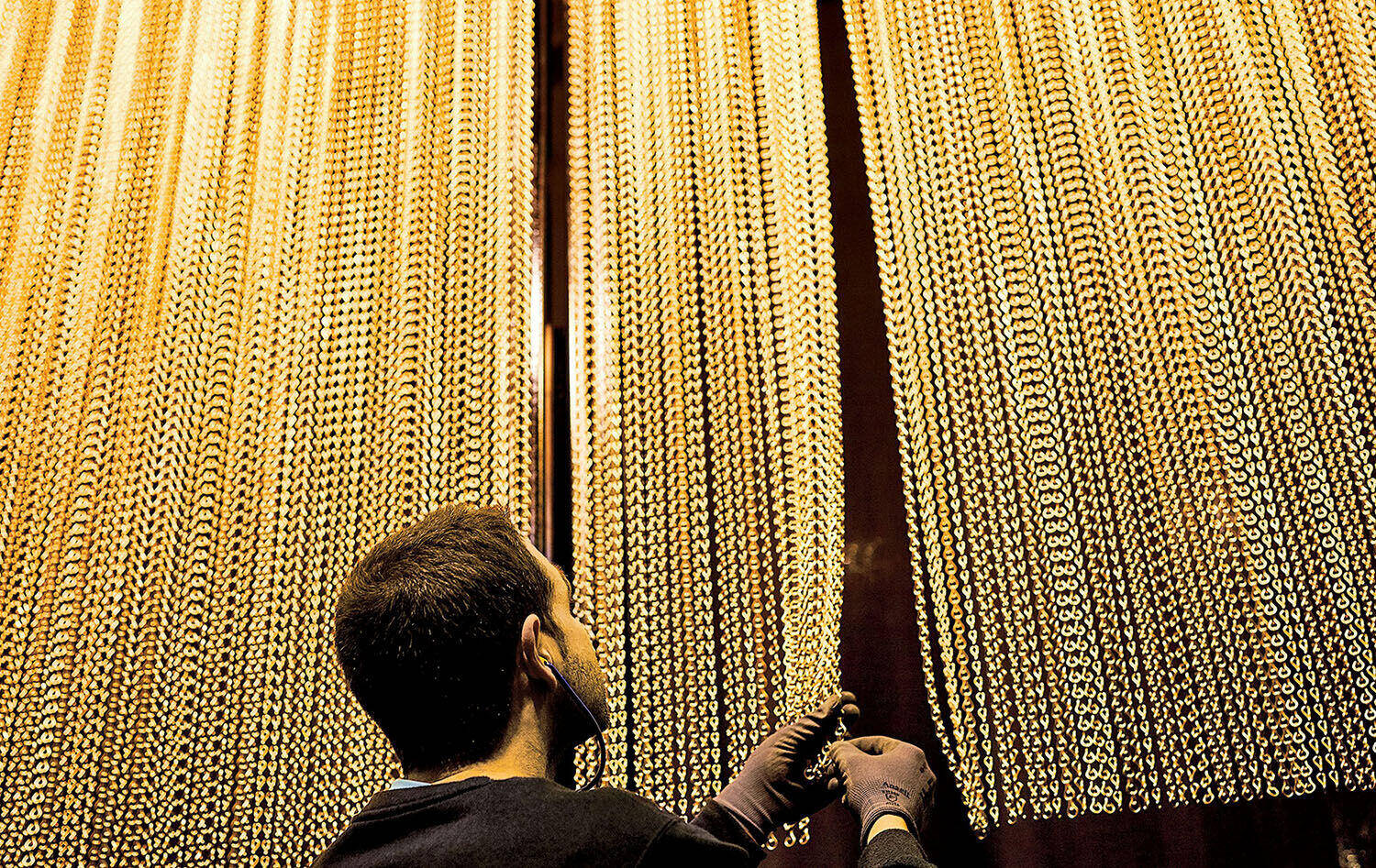 A person wearing gloves inspects and arranges long gold chains hanging in parallel rows, facing tall, brightly lit displays.