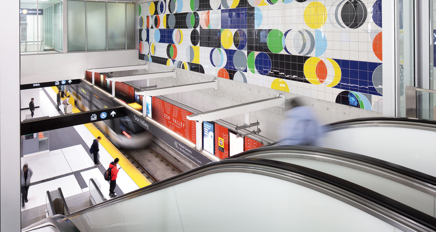 People wait on a subway platform as a train arrives; colorful geometric tile art decorates the wall above the platform, and escalators are in the foreground.
