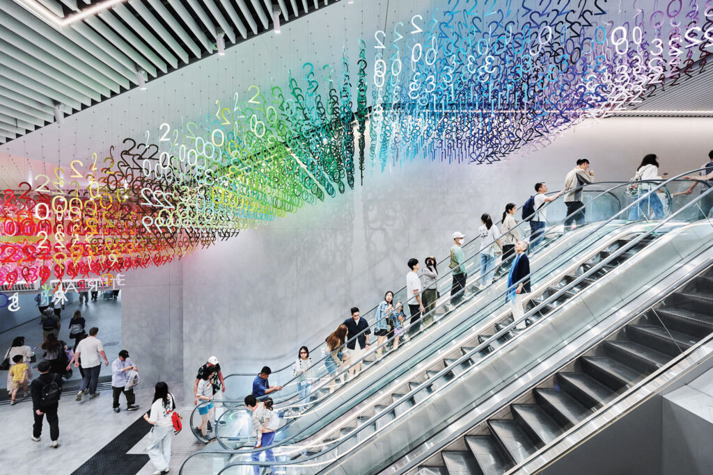 People ride escalators in a modern building with colorful numbers hanging from the ceiling in a rainbow gradient.