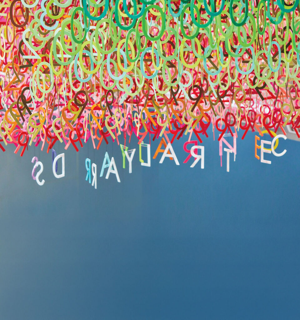 Colorful plastic letters hang densely from the ceiling, with a few letters spaced out against a blue background, appearing upside down.