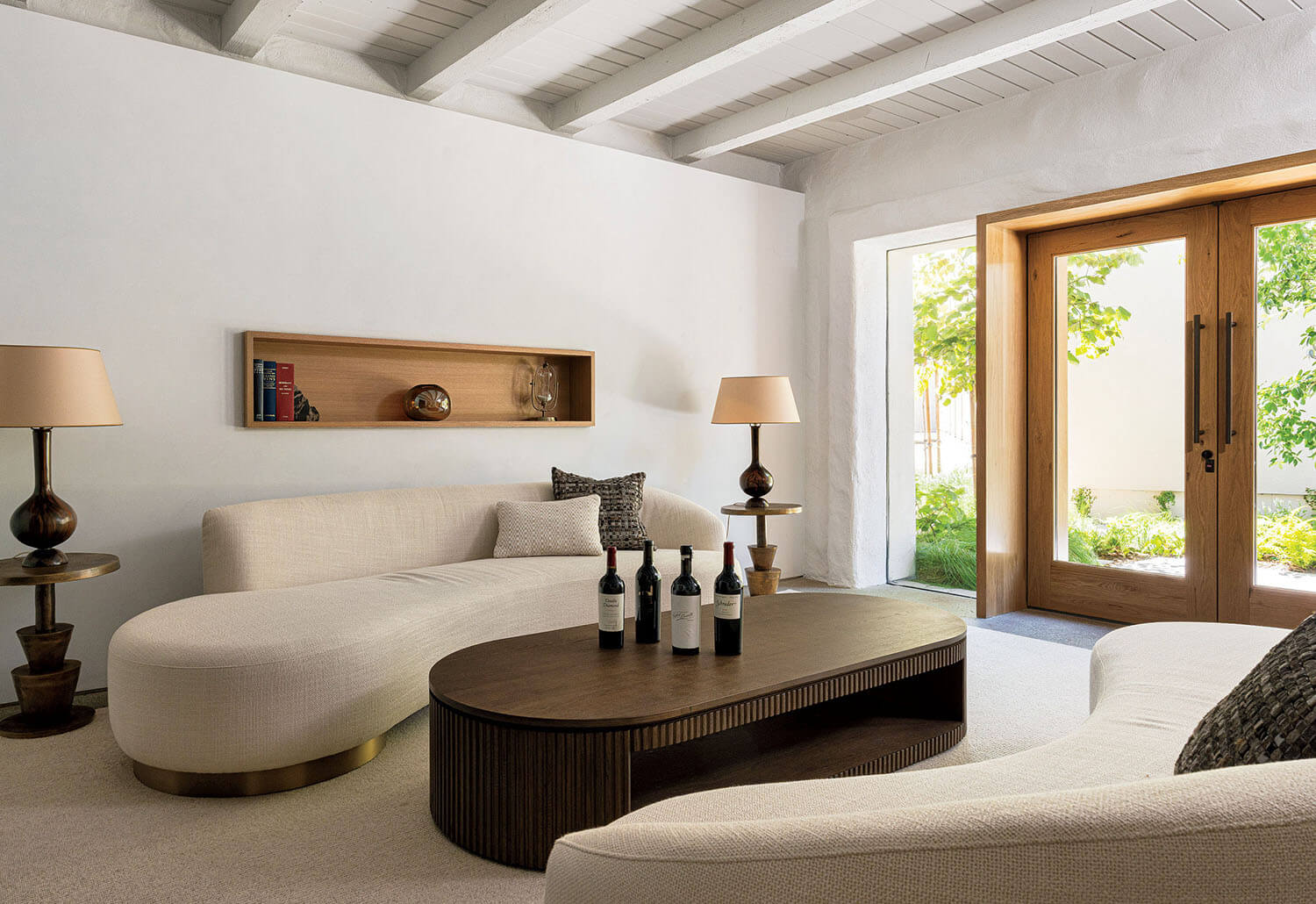 Curved cream sofas and a large wooden coffee table with four wine bottles in a brightly lit living room.