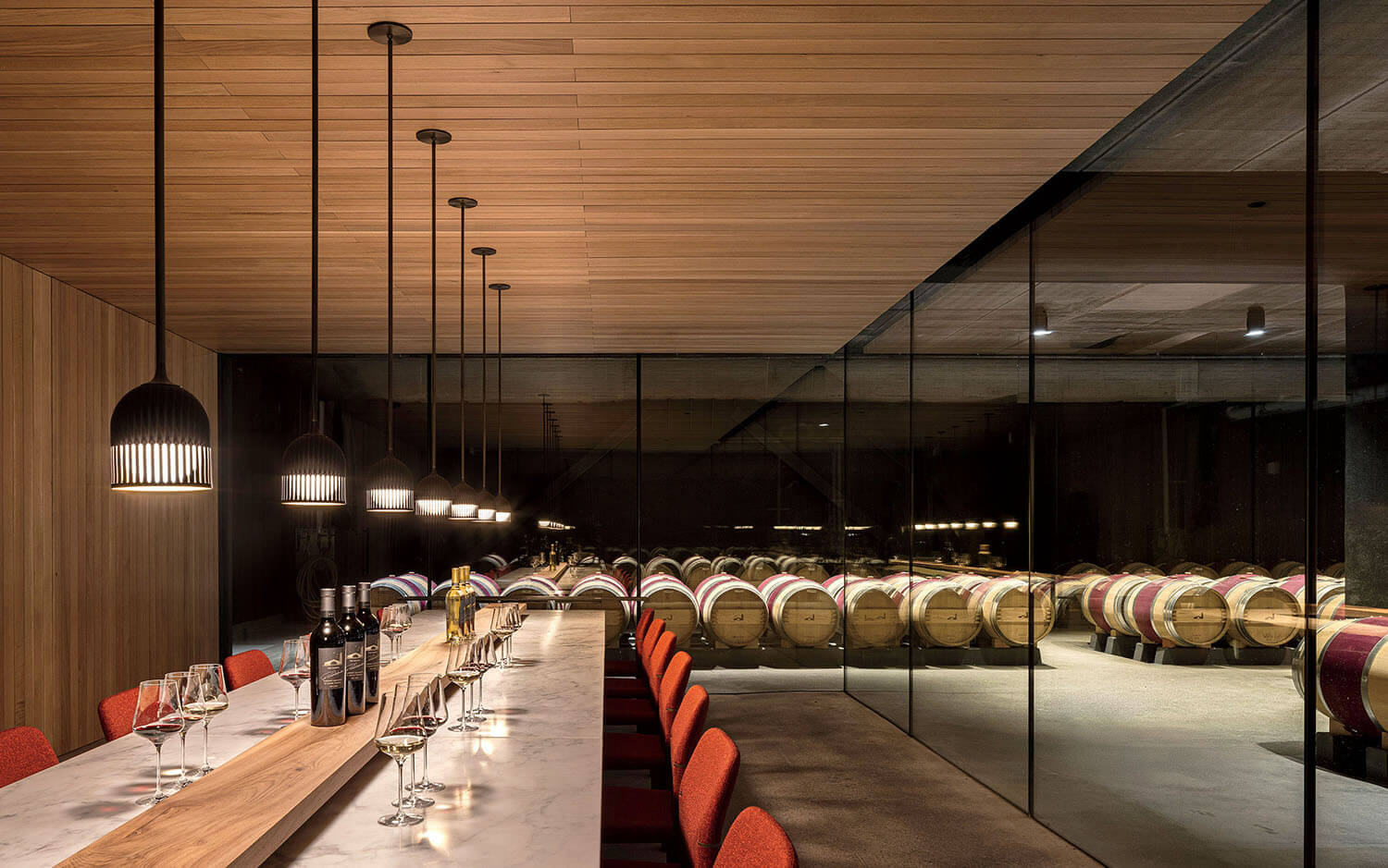 A long table with wine glasses and bottles overlooks a cellar filled with stacked wooden wine barrels behind a glass partition.