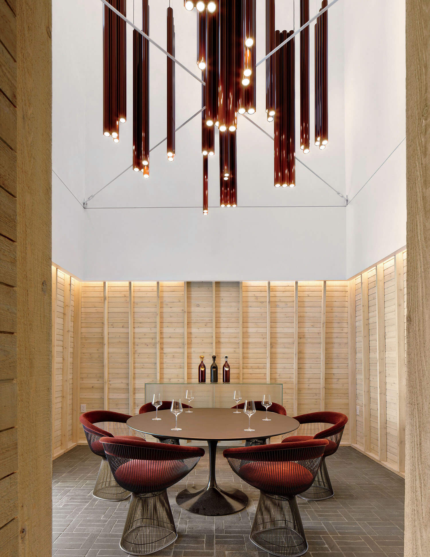 A circular dining table with four red chairs, set under a large, tiered chandelier made of cylindrical glass lights, in a room with wood-paneled walls.