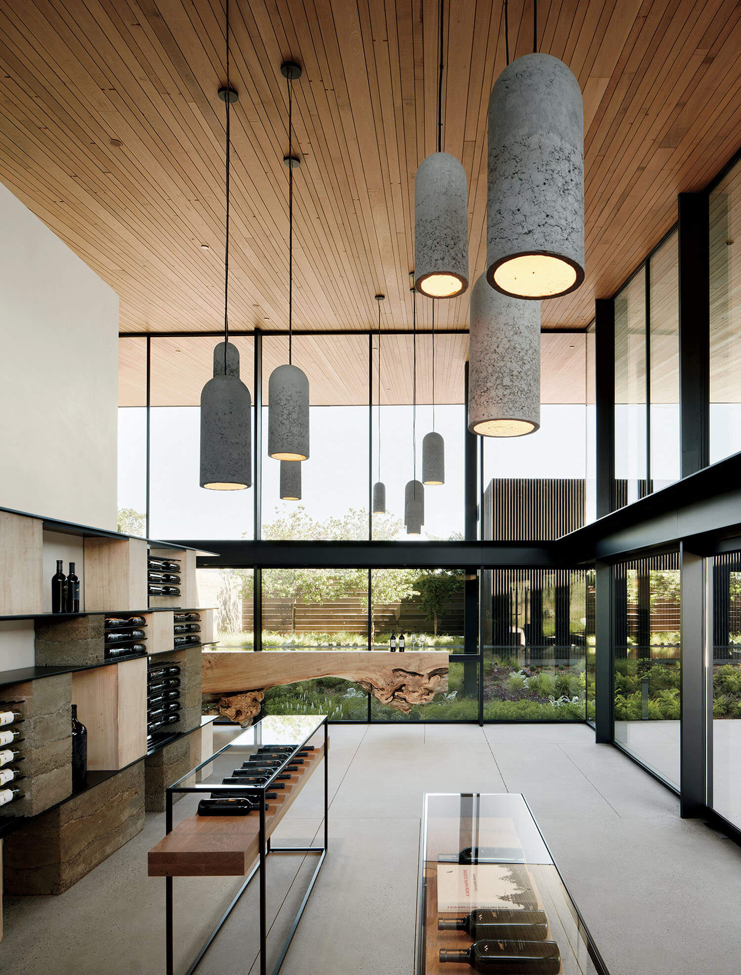 Modern room with tall windows, large cylindrical concrete pendant lights, and wooden shelving displaying wine bottles.