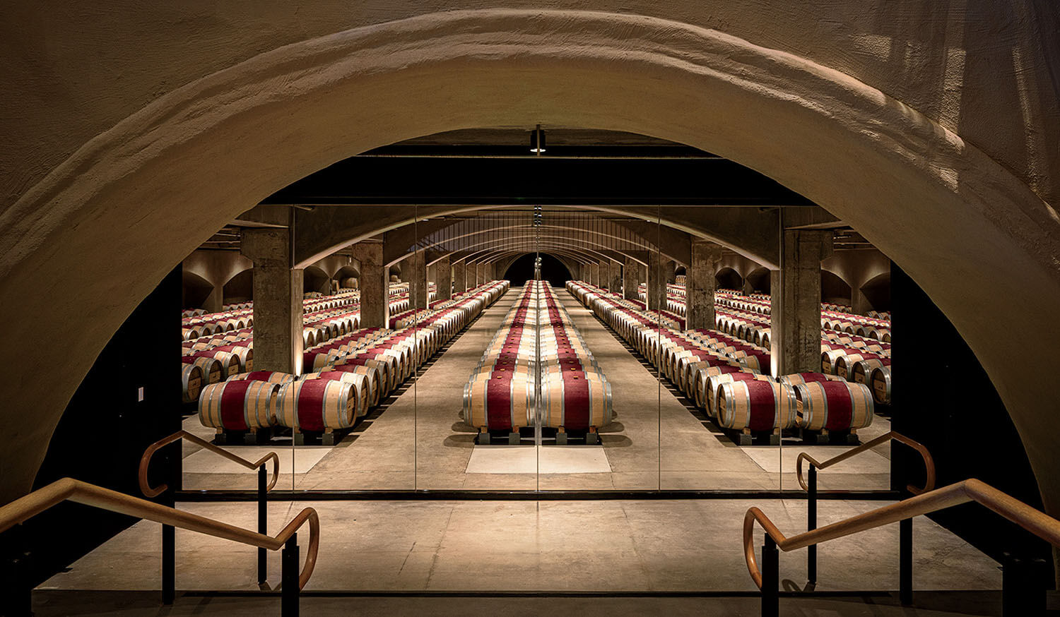 A long, symmetrical view of a wine cellar with rows of wooden barrels under a stone arched ceiling, seen through a glass partition.