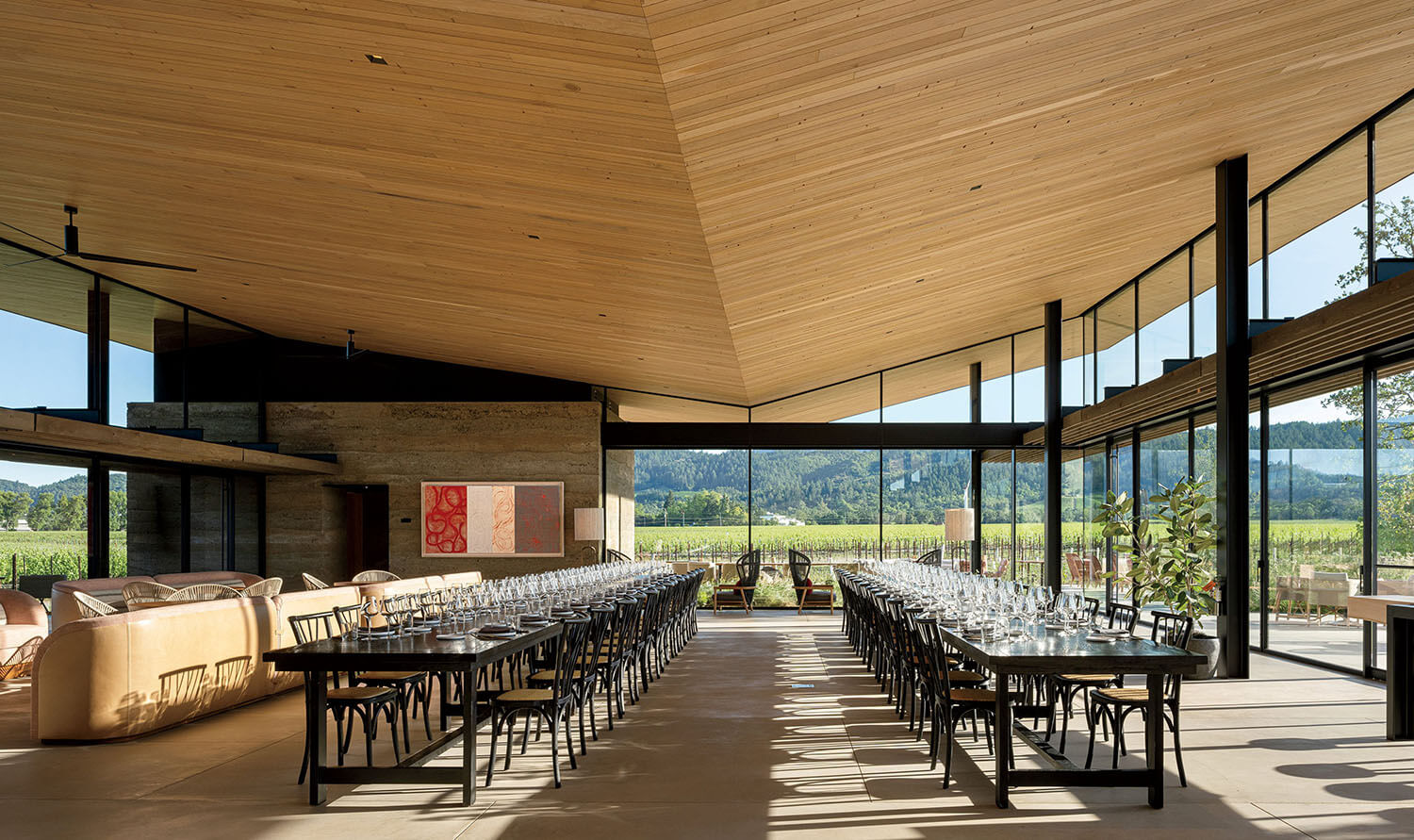 A modern dining room with two long tables set for dinner, featuring floor-to-ceiling windows overlooking a vineyard.