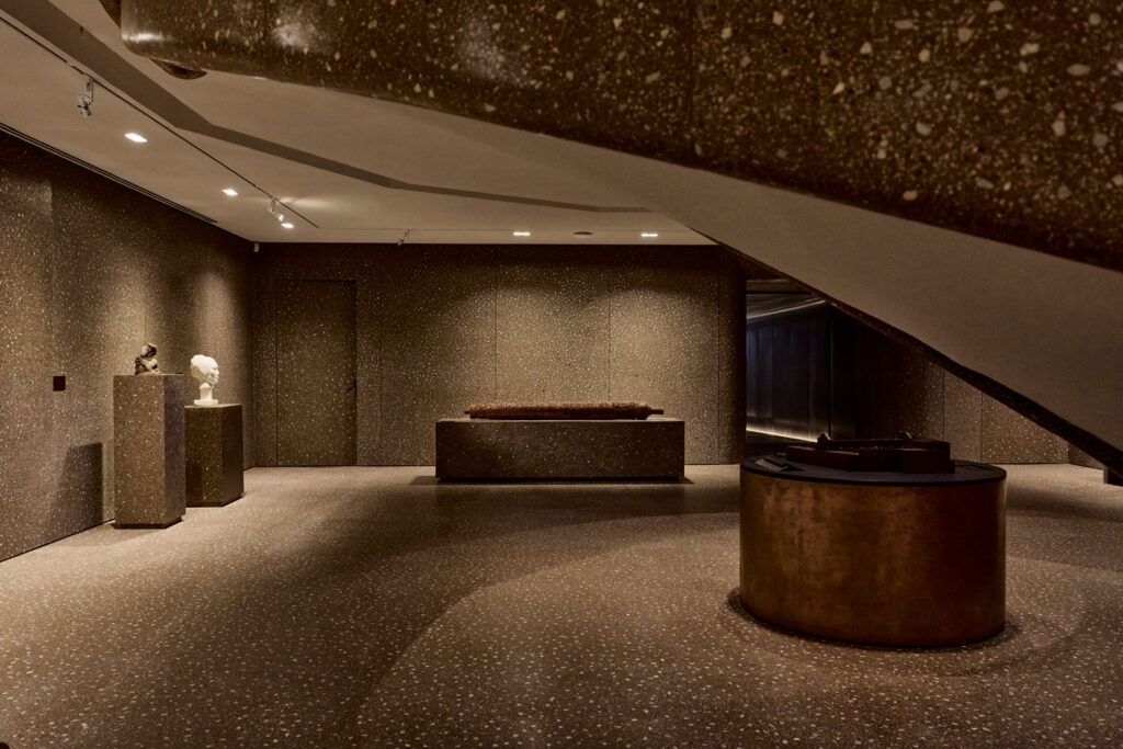 Gallery interior with speckled stone walls and flooring, featuring sculptural pedestals, a long display platform, and a circular bronze base beneath a curved staircase.