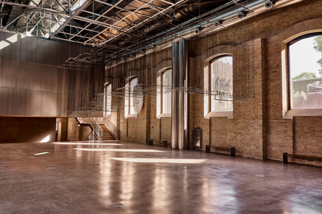 Large, empty industrial-style hall with exposed rafters, brick walls, arched windows, and a polished concrete floor.