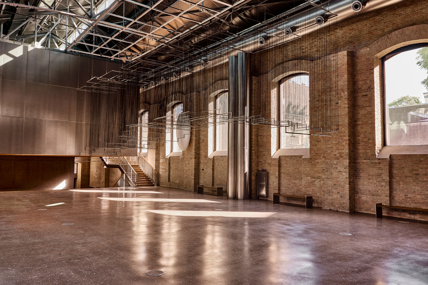 Large, empty industrial-style hall with exposed rafters, brick walls, arched windows, and a polished concrete floor.