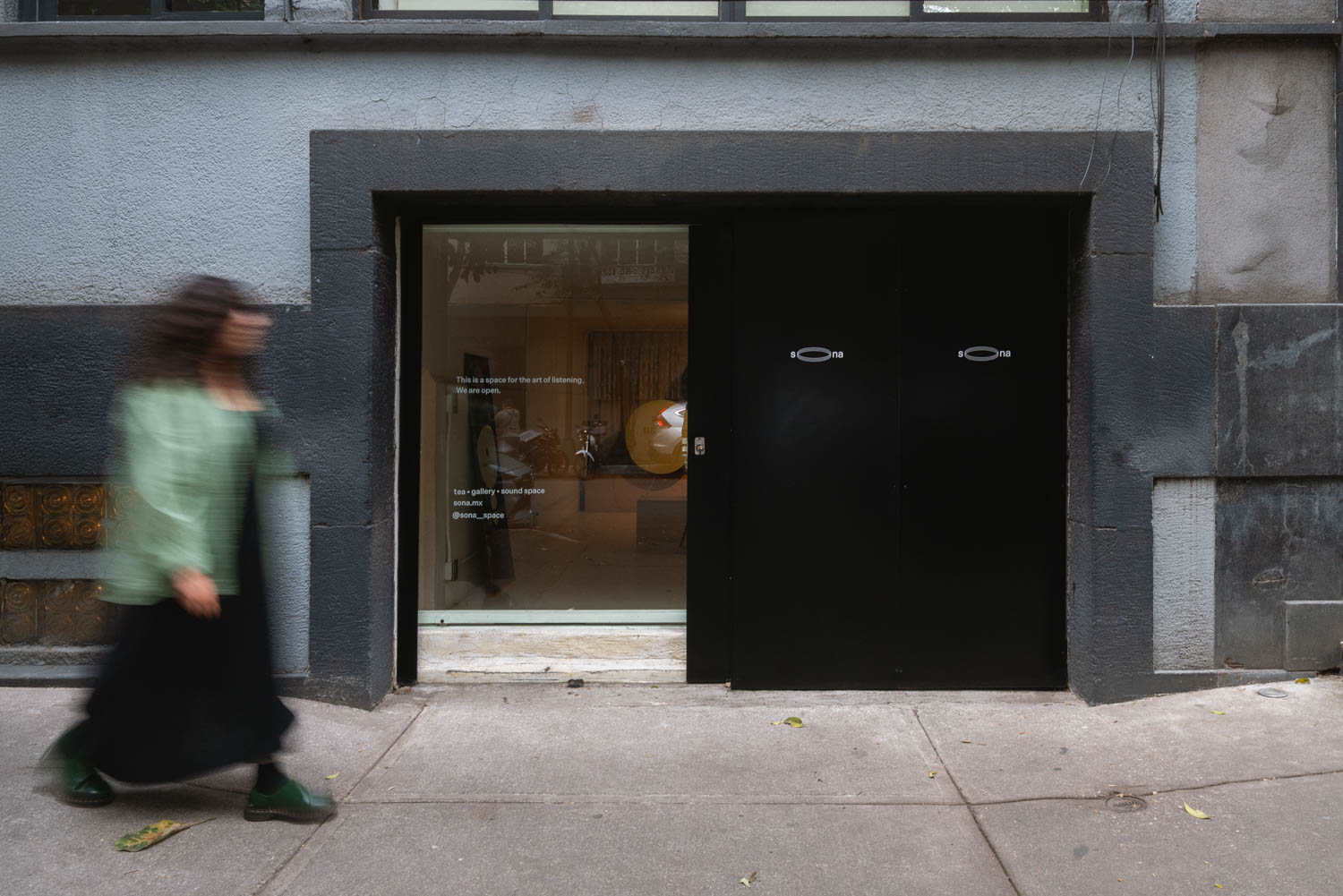 A person walks past the entrance of Sona, a tea and sound space with a storefront window featuring the business name and information.