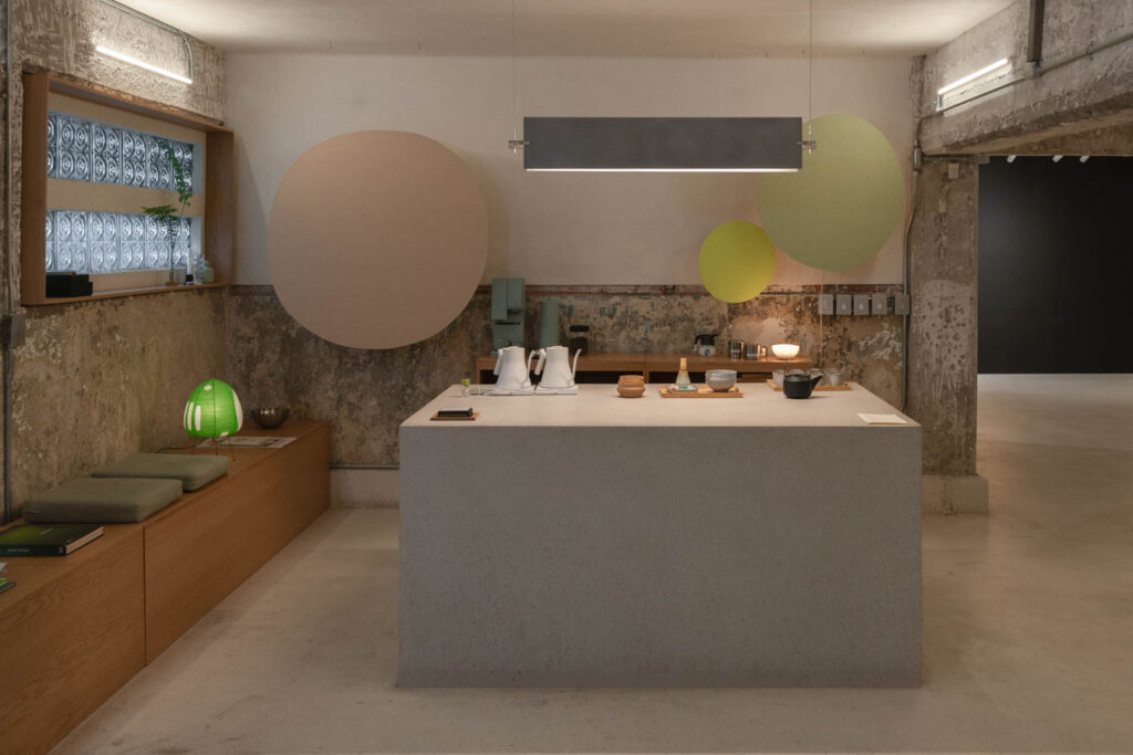 A kitchen counter with tea equipment, placed in a room with a concrete island, bench seating, and circular wall art.