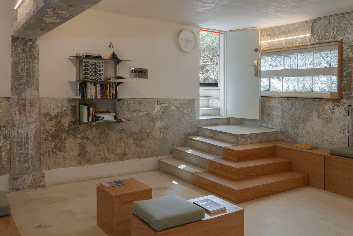 A minimalist room with a concrete wall, wooden platform steps, a wall-mounted bookshelf, and a doorway leading to a small outdoor staircase.
