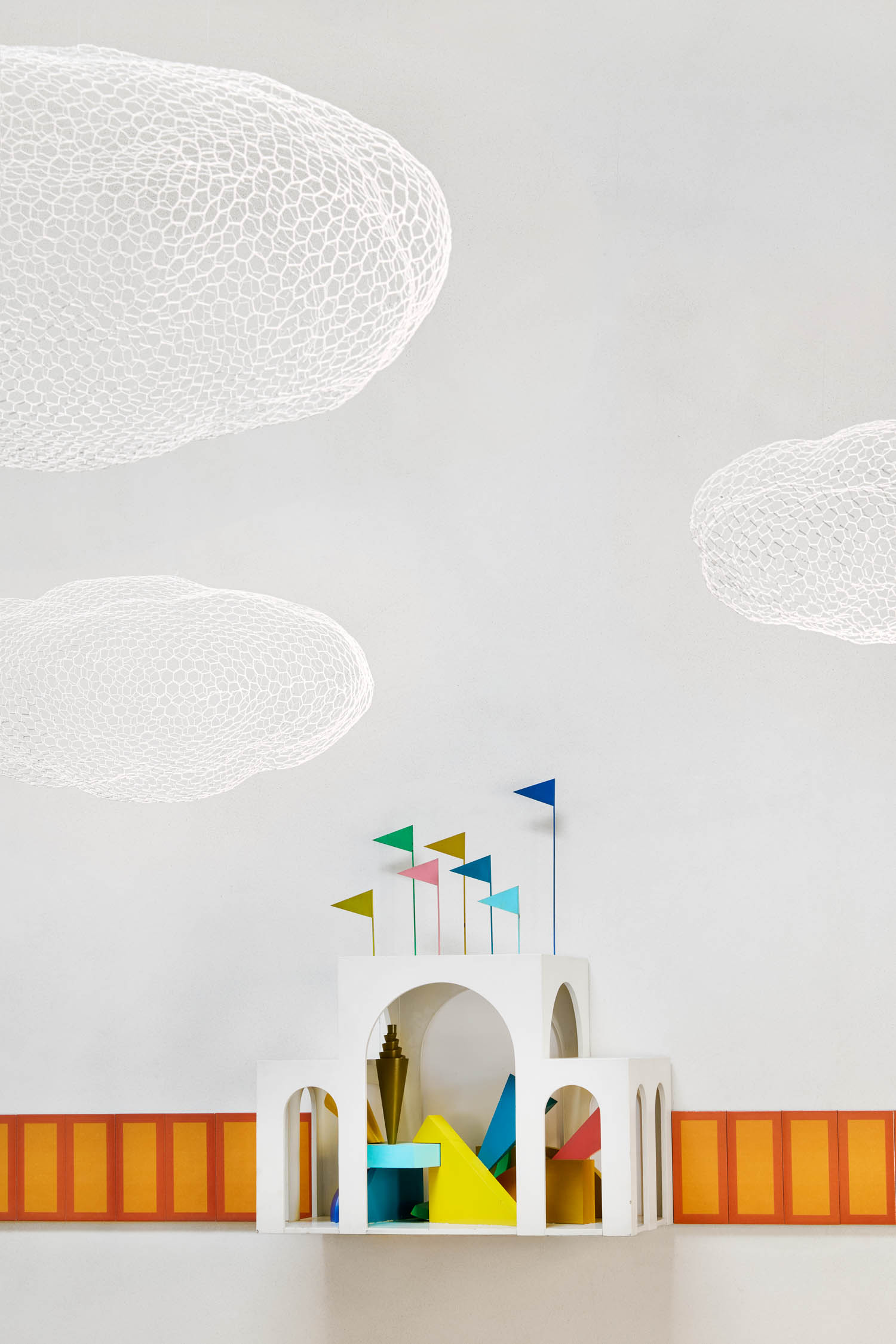 A white architectural structure with colorful geometric shapes and flags, set against a wall with orange panels and suspended wire-mesh clouds.