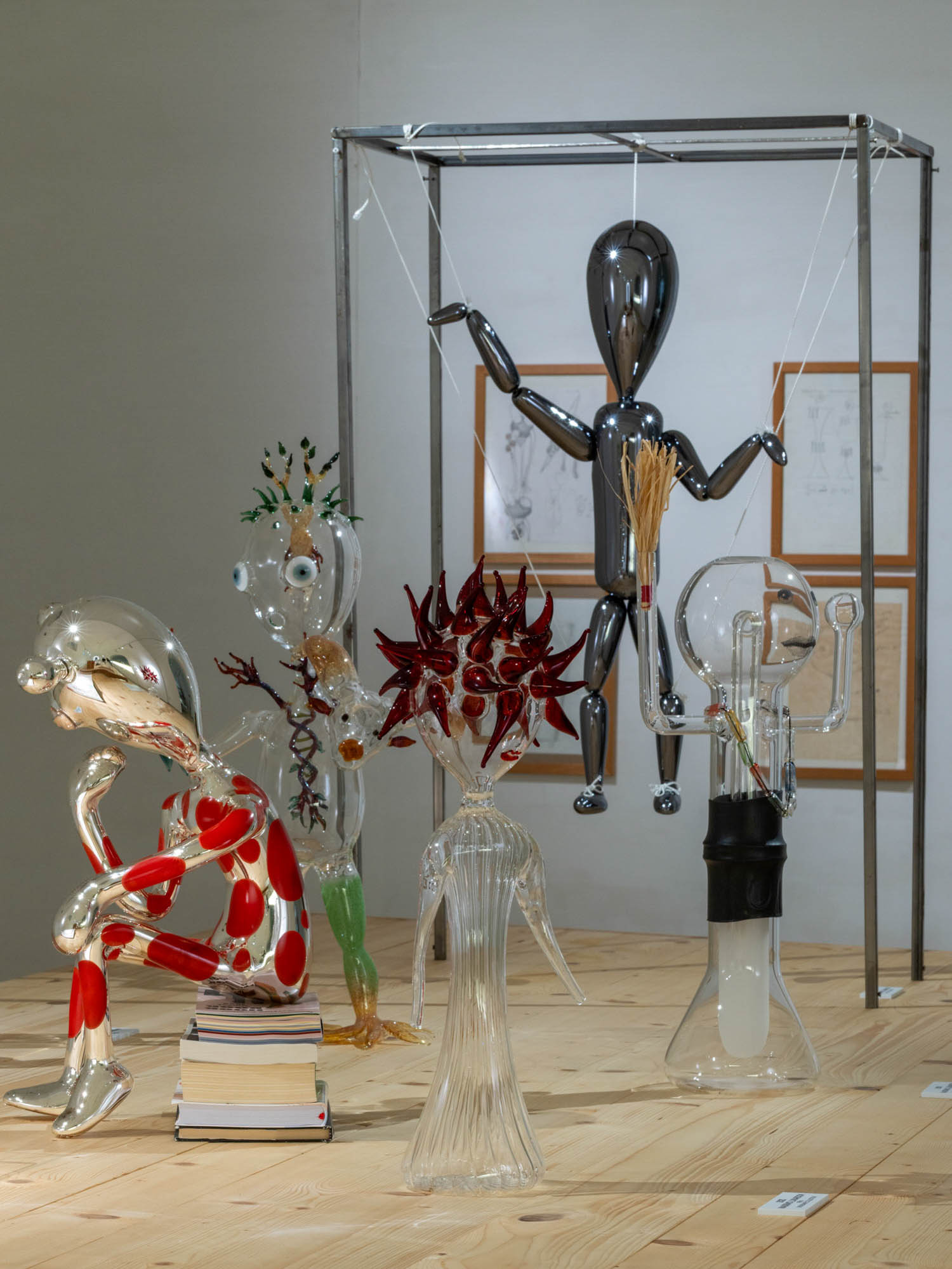 Five glass sculptures of humanoid figures on a wooden floor, with one chrome figure suspended from a metal frame.